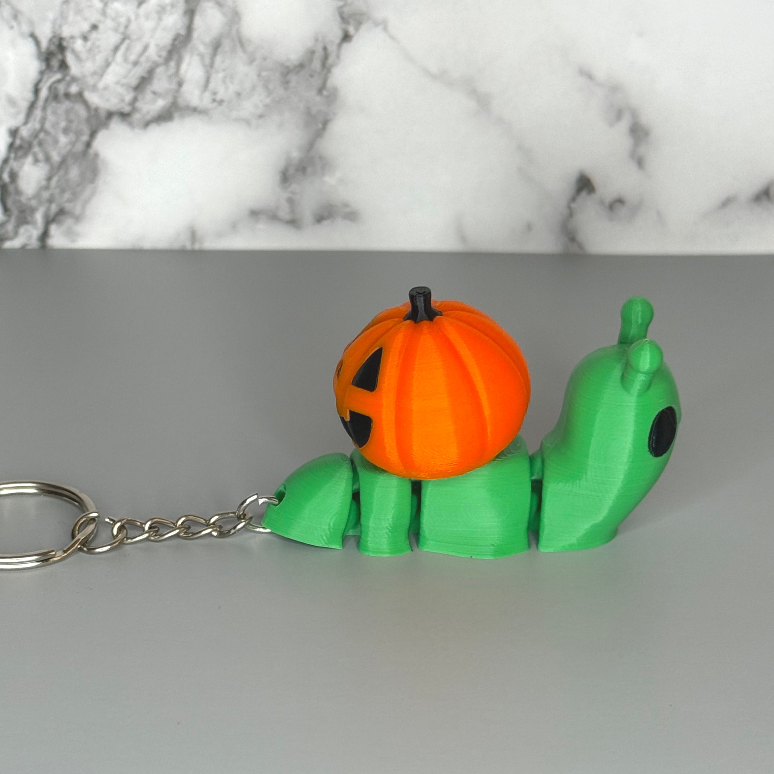 Side of green snail-shaped keychain with orange jack-o-lantern shell on a gray background.