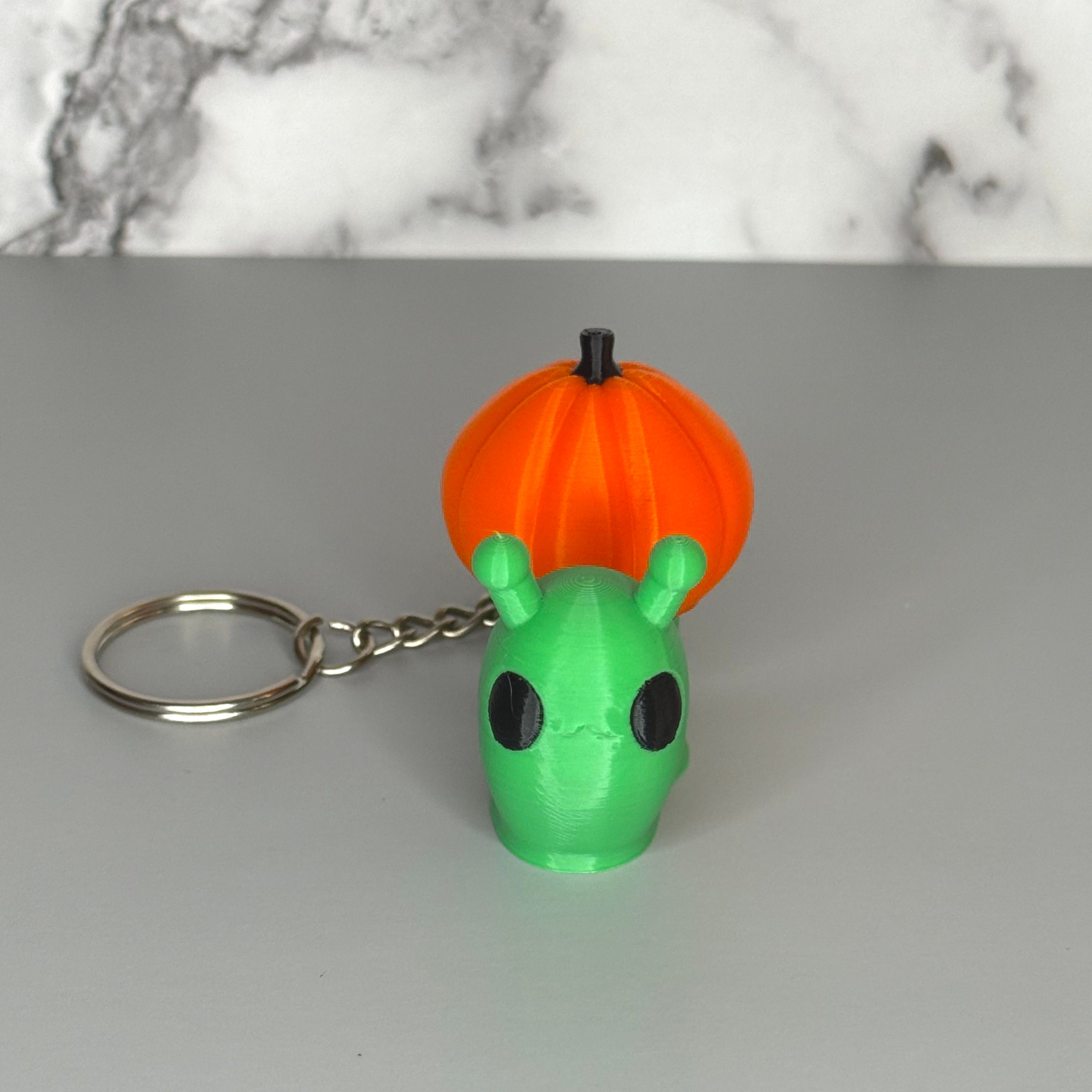 Green snail-shaped keychain with orange pumpkin shells on a gray marble background.