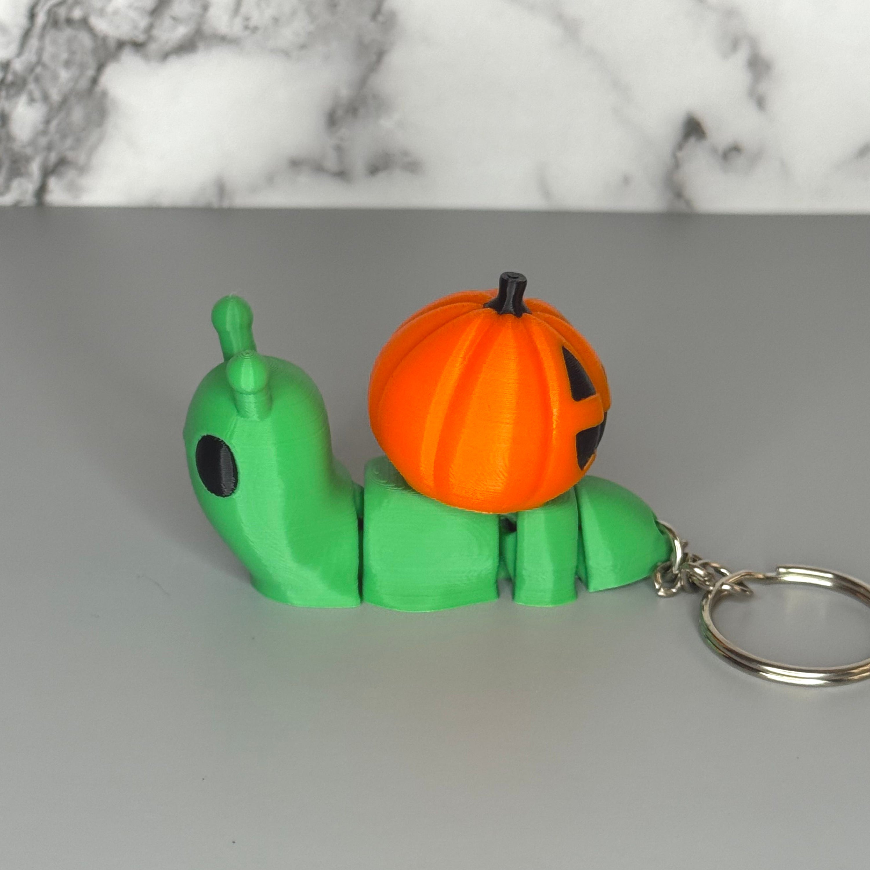 Side of green snail-shaped keychain with orange jack-o-lantern shell on a gray background.
