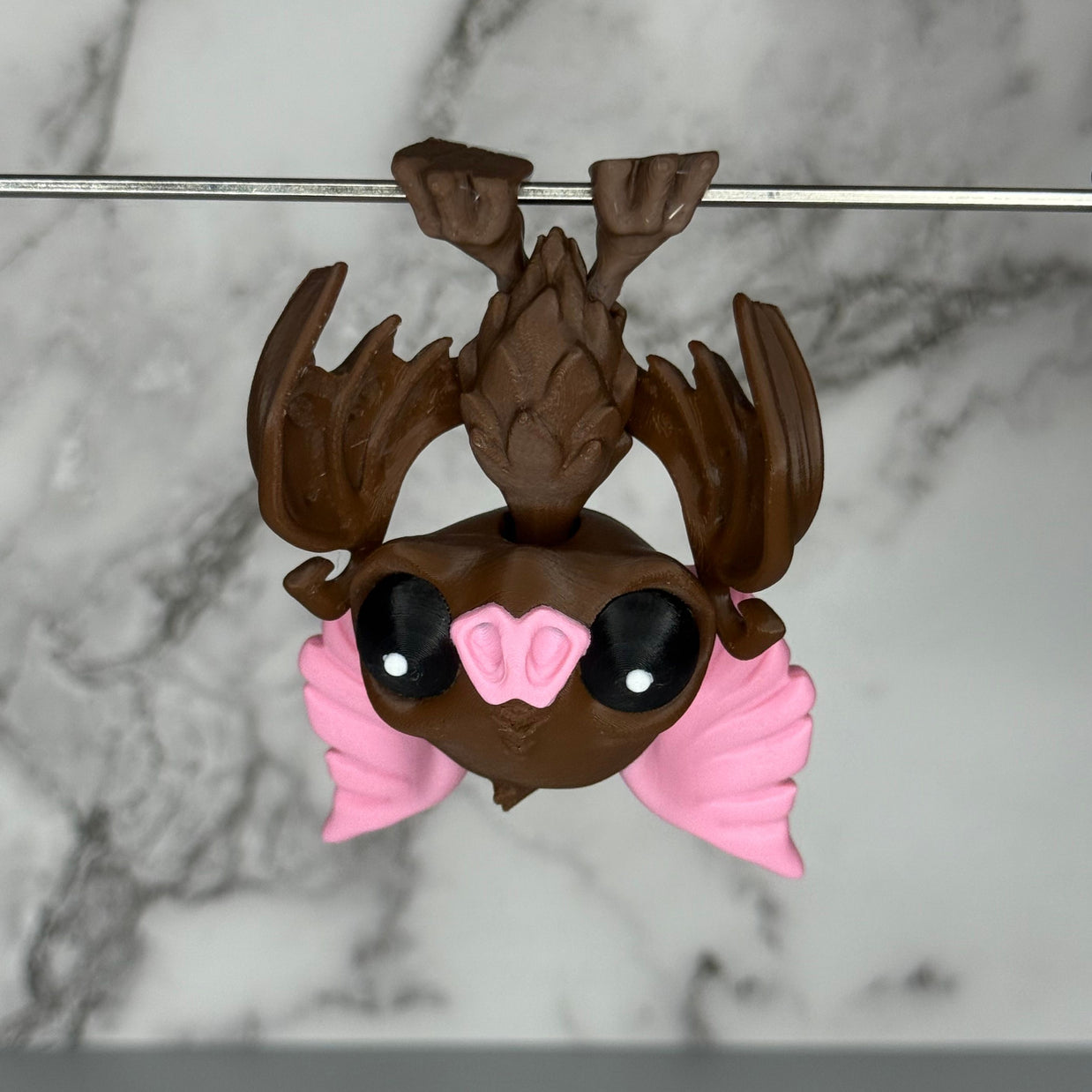 Toy figure of a brown bat with pink ears and nose hanging from a silver rod on a marble gray background