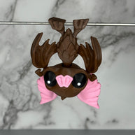 Toy figure of a brown bat with pink ears and nose hanging from a silver rod on a marble gray background