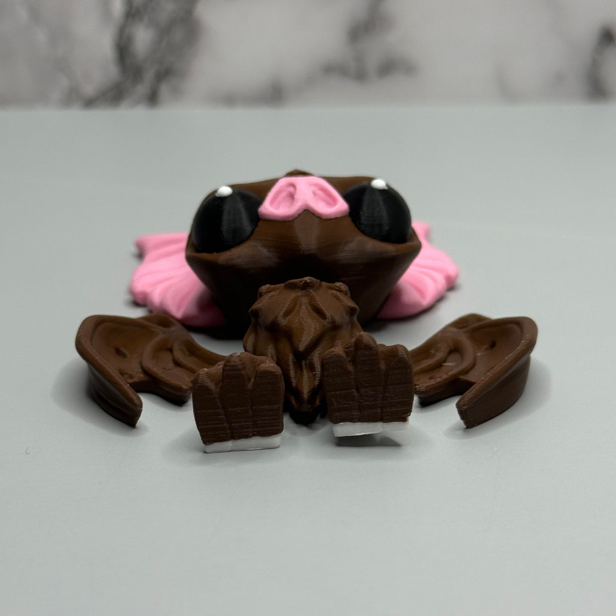 Bottom side of a figure of a brown bat with pink ears and nose on a gray and marble gray background