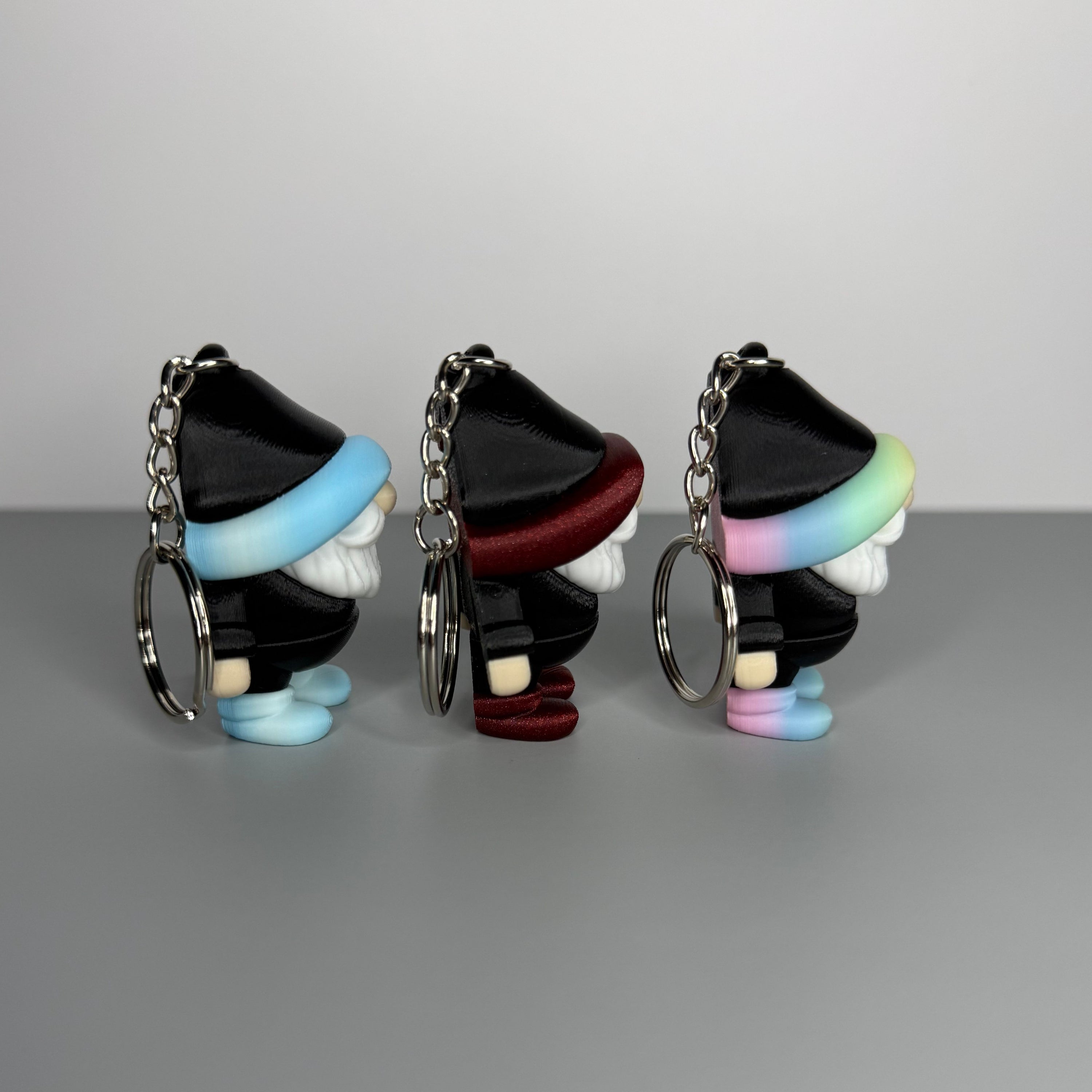 Left side of three gnome-shaped keychains in black with different colored hats (blue/white gradient and black, shimmer red and black, and pastel rainbow gradient and black) with shoes the same color as the hats on a gray background