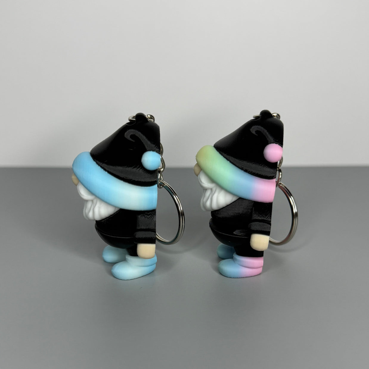 Right side of two gnome-shaped keychains in black with different colored hats (blue/white gradient and black, and pastel rainbow gradient and black) with shoes the same color as the hats on a gray background