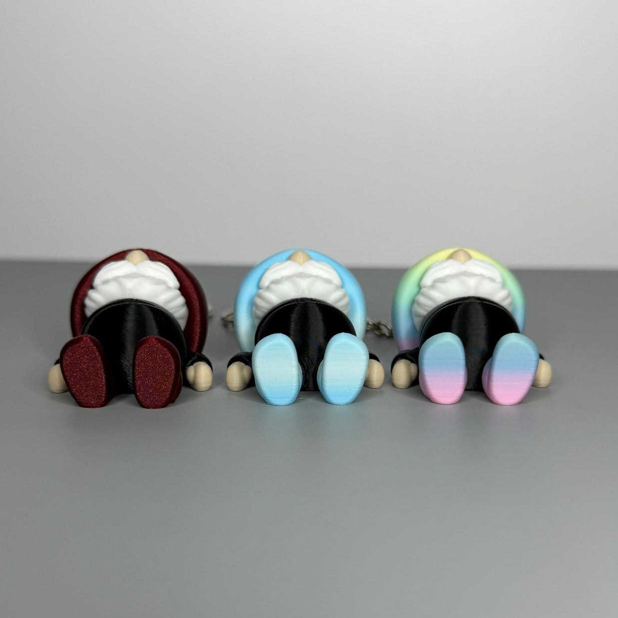 Bottom side of three gnome-shaped keychains in black with different colored hats (blue/white gradient and black, shimmer red and black, and pastel rainbow gradient and black) with shoes the same color as the hats on a gray background