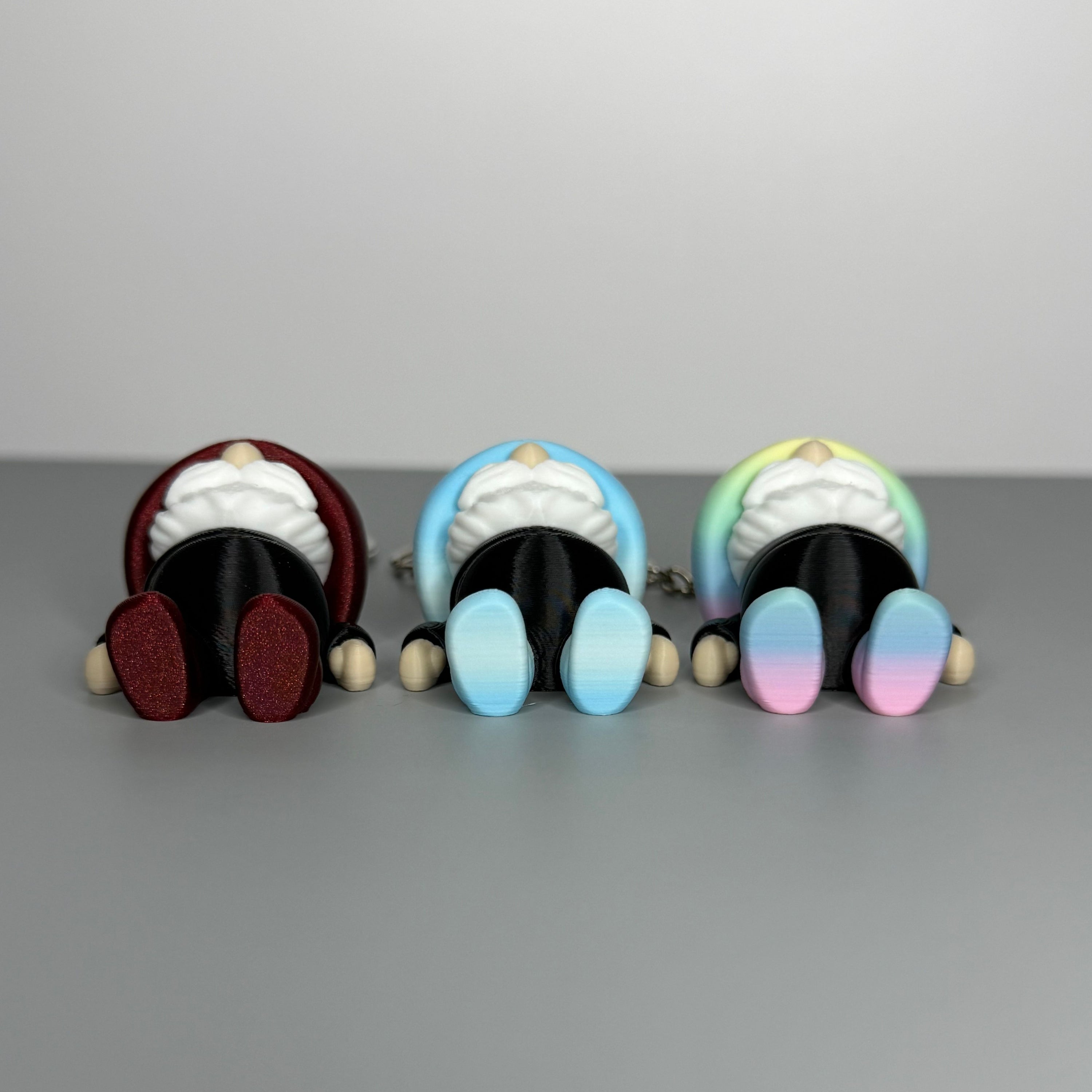 Bottom side of three gnome-shaped keychains in black with different colored hats (blue/white gradient and black, shimmer red and black, and pastel rainbow gradient and black) with shoes the same color as the hats on a gray background