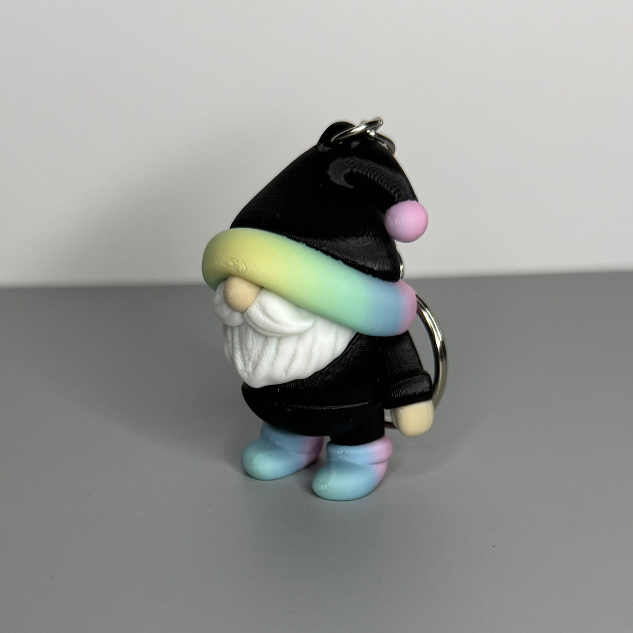 Gnome-shaped keychain in black with a pastel rainbow and black hat, white beard, and pastel rainbow shoes. 