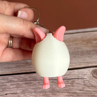 Hand holding a 3D printed white egg with pink bunny ears and legs keychain on a wooden surface