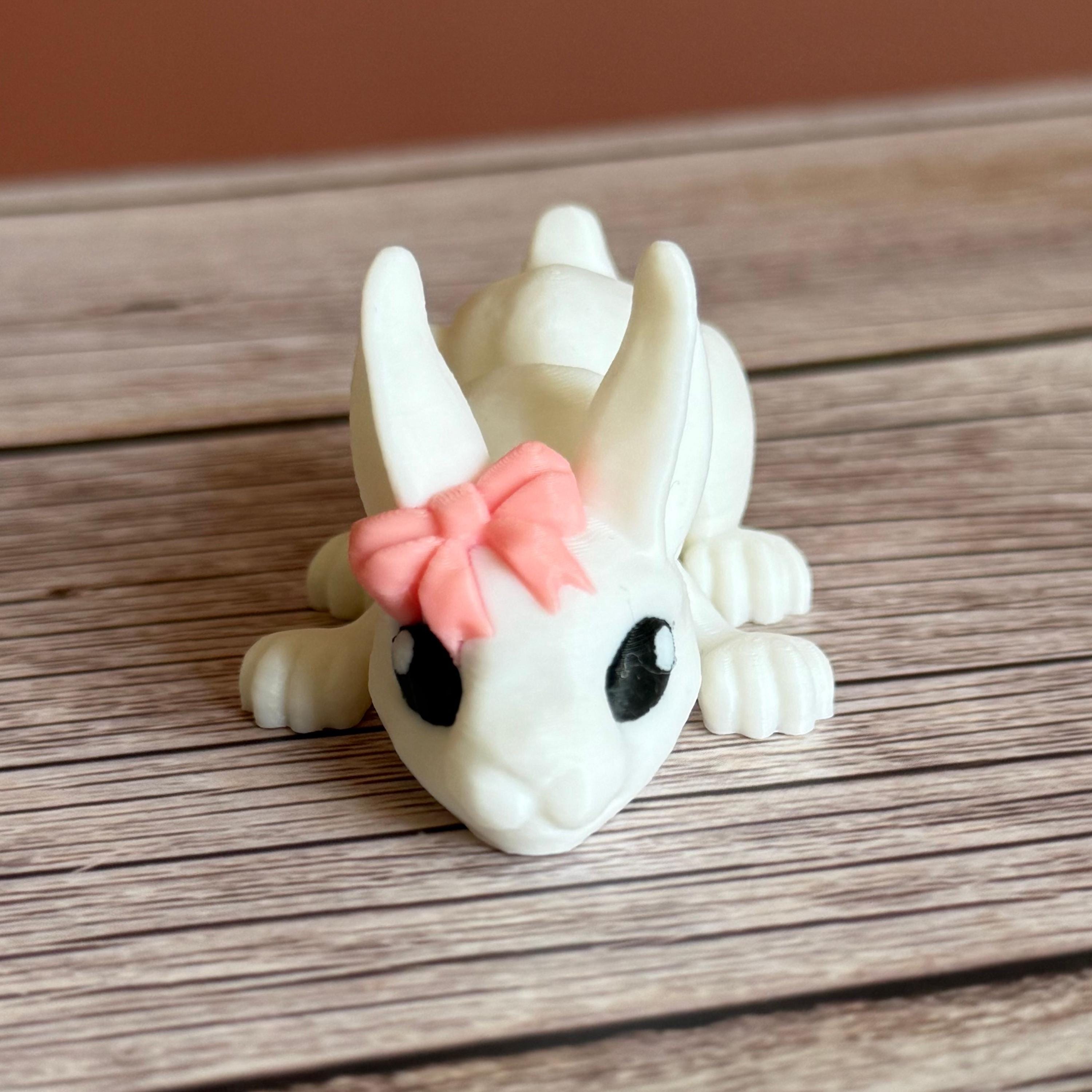 Front of a 3D printed white rabbit figurine with a pink bow on a wooden surface
