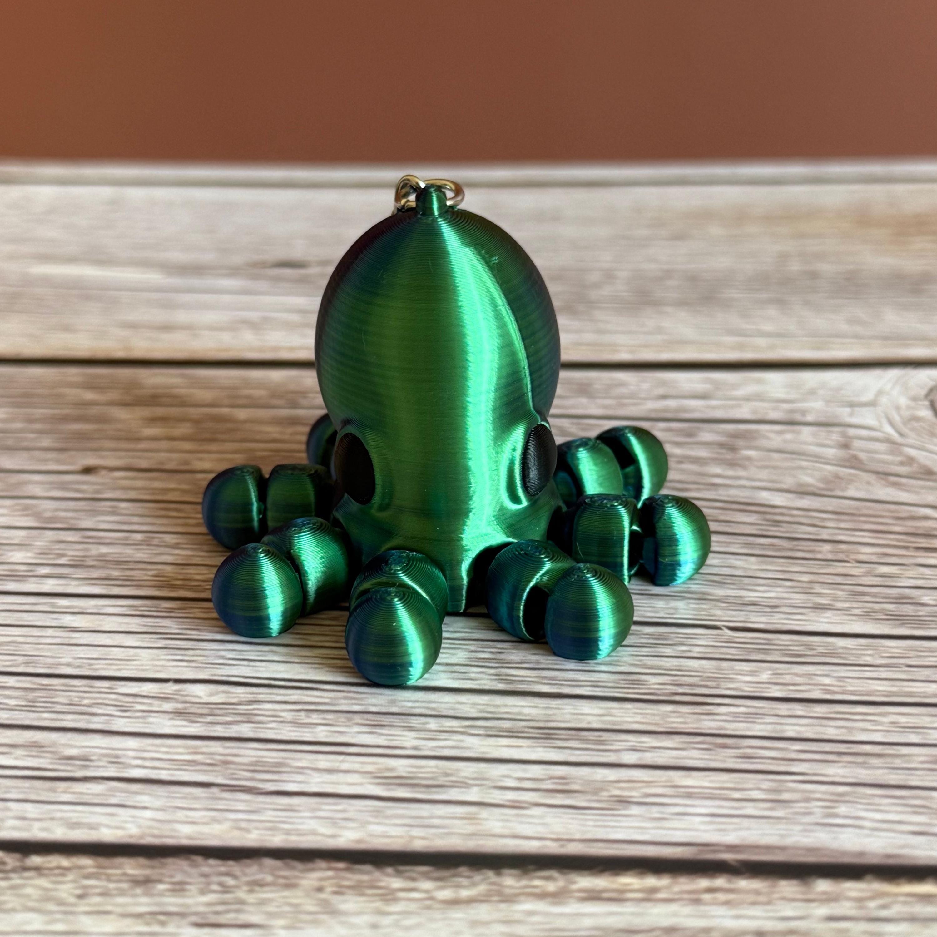 3D printed shiny green and blue octopus-shaped keychain, placed on a wooden surface with a brown background