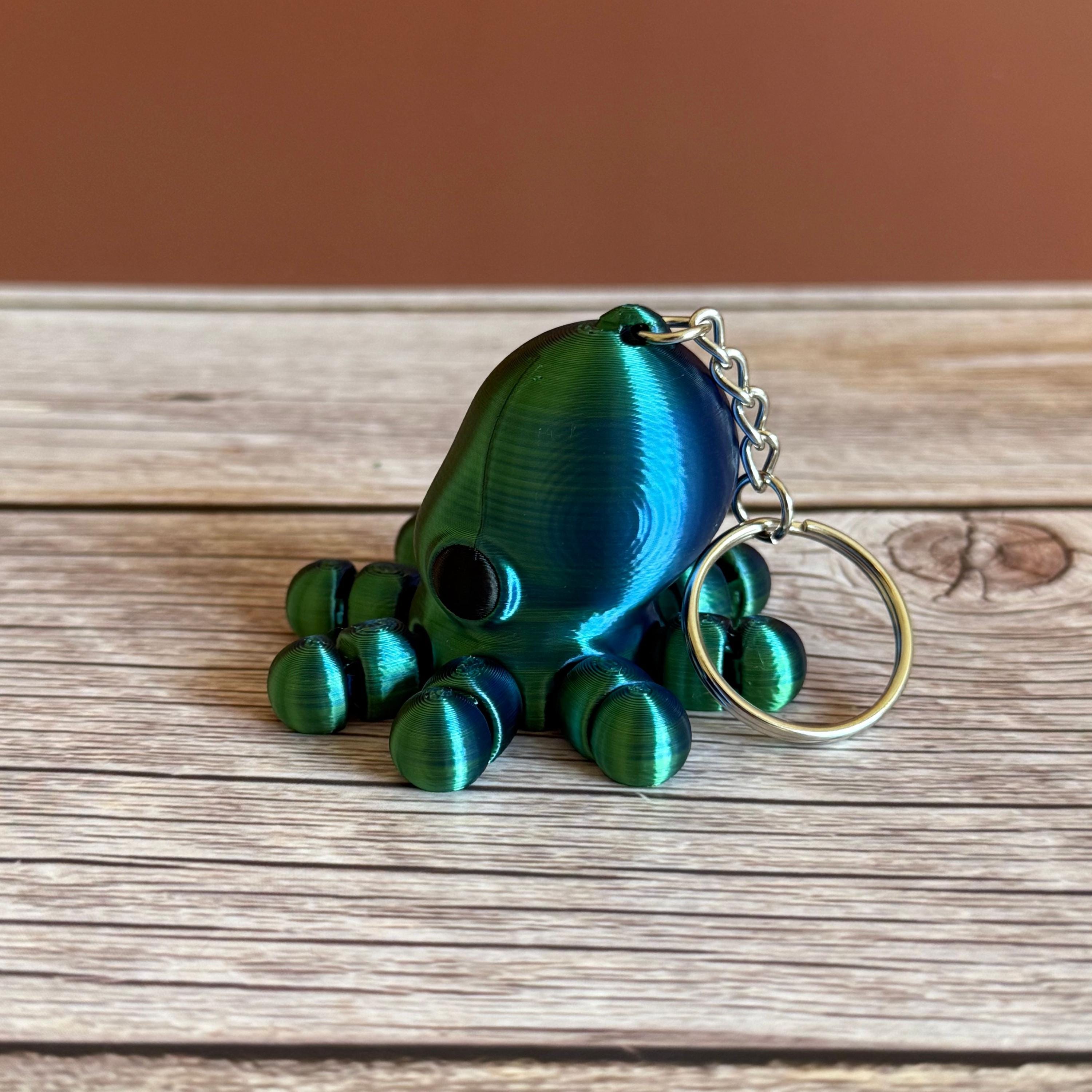 3D printed shiny green and blue octopus-shaped keychain, placed on a wooden surface with a brown background