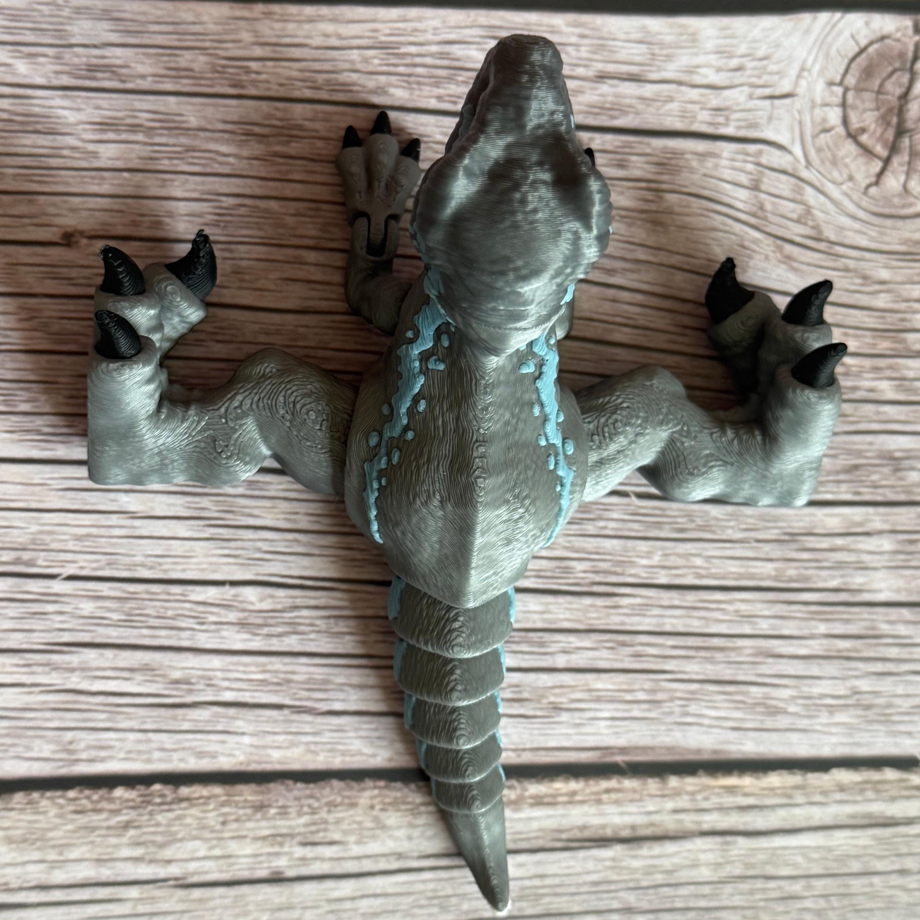 Top of a 3D printed blue, gray, and white colored raptor dinosaur figure with legs spread wide, placed on a wooden surface against a brown background