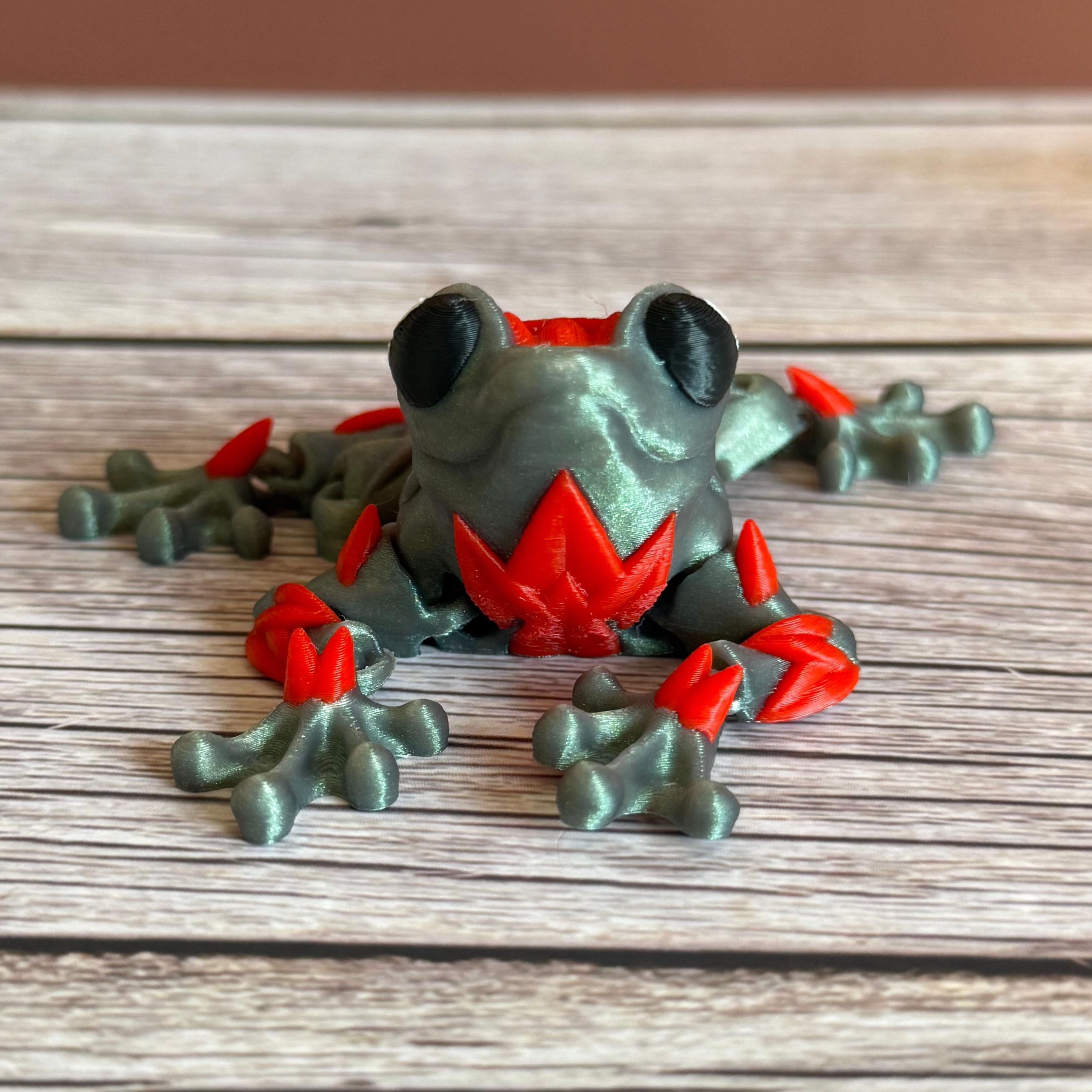 3D printed shimmer-green frog with red rose on it's back, placed on a wooden surface against a brown background