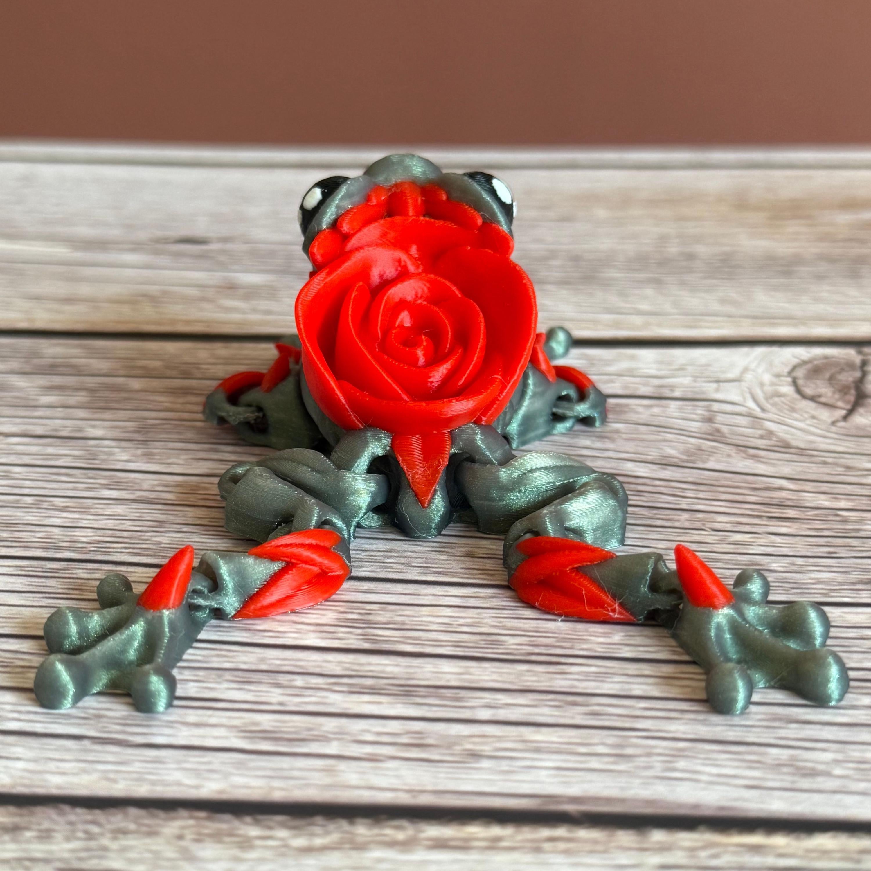 Back of a 3D printed shimmer-green frog with red rose on it's back, placed on a wooden surface against a brown background