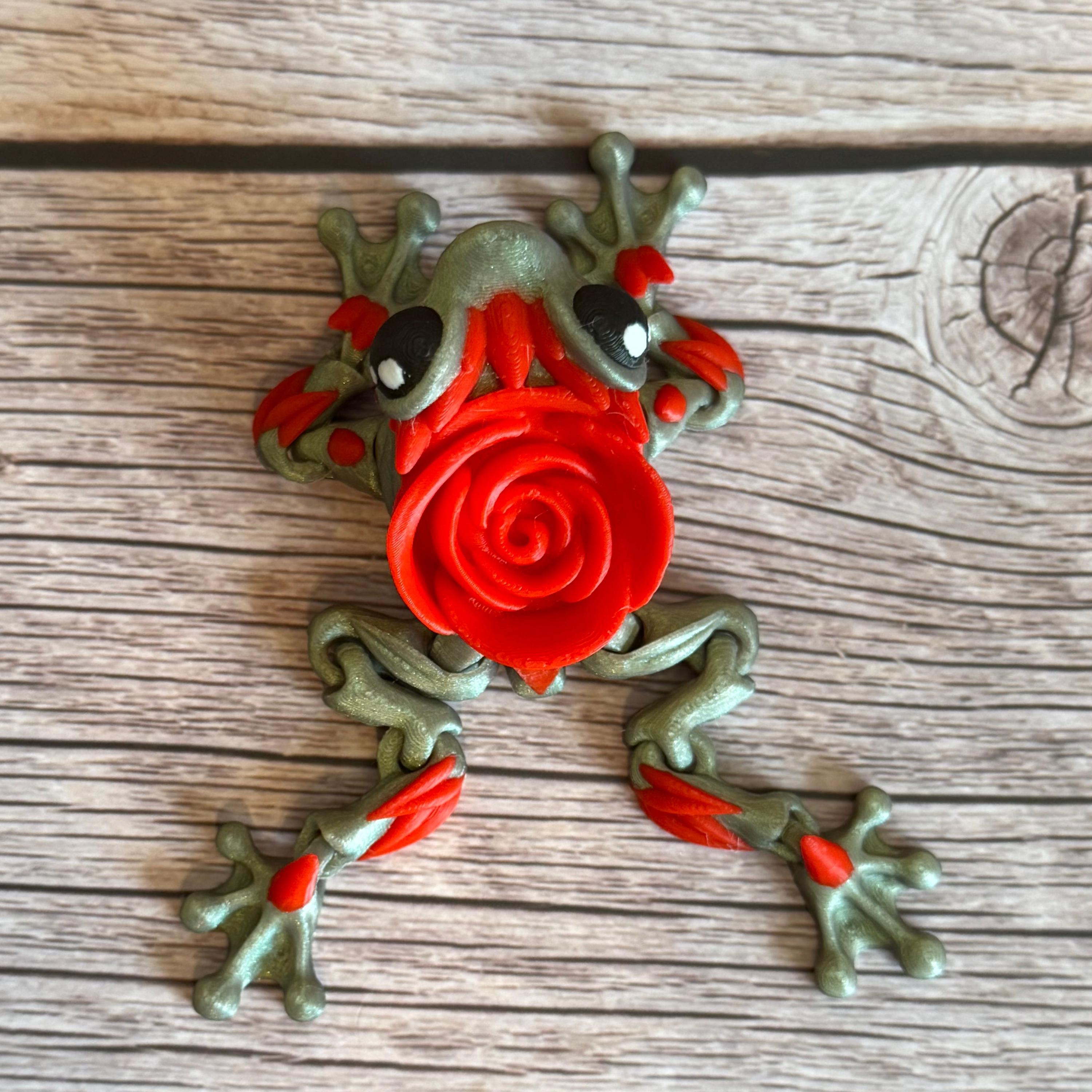 Top of a 3D printed shimmer-green frog with red rose on it's back, placed on a wooden surface