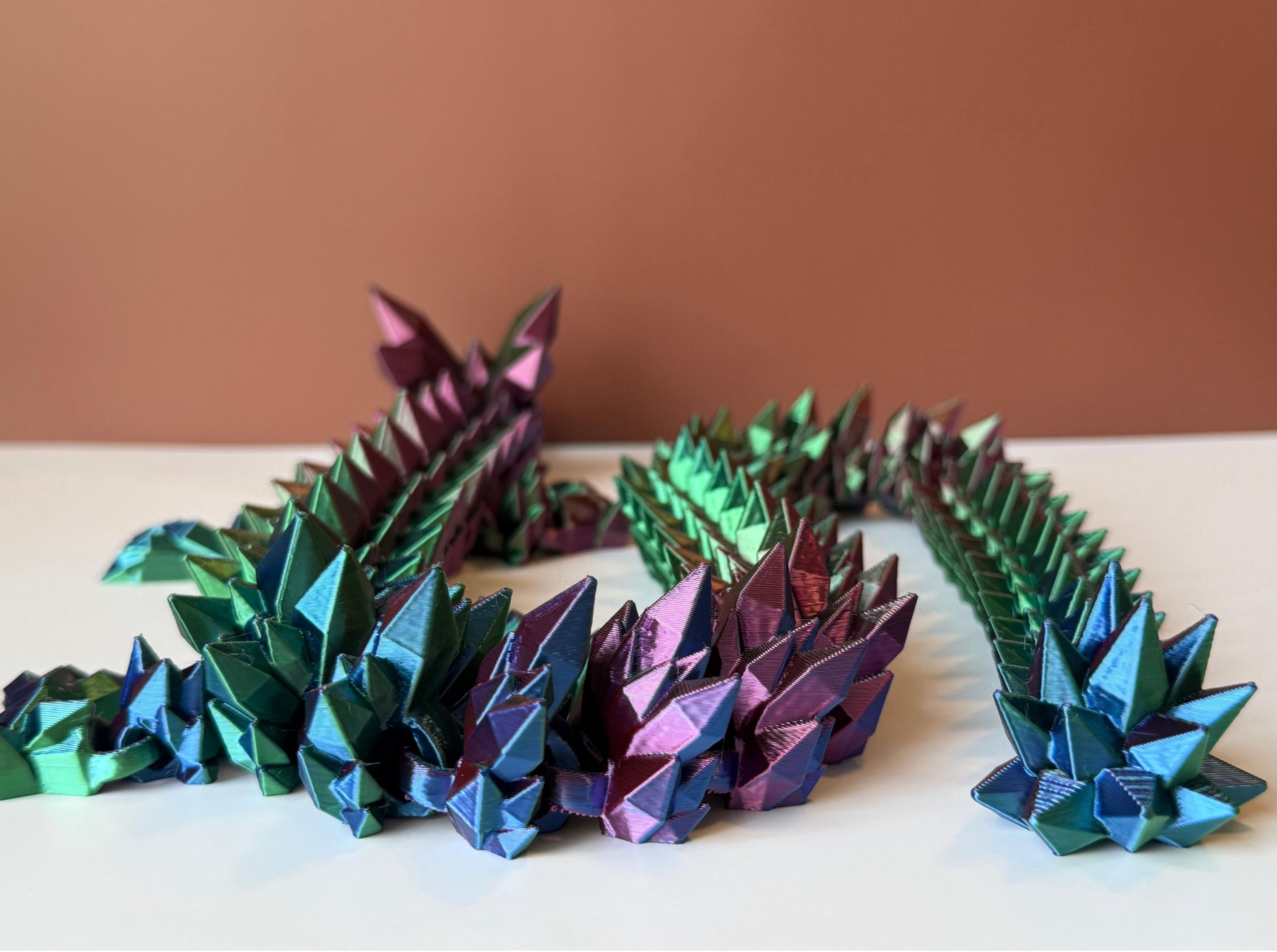 Multicolored 3D printed dragon with crystal-shaped features across the entire body, placed on a white surface with a brown backdrop