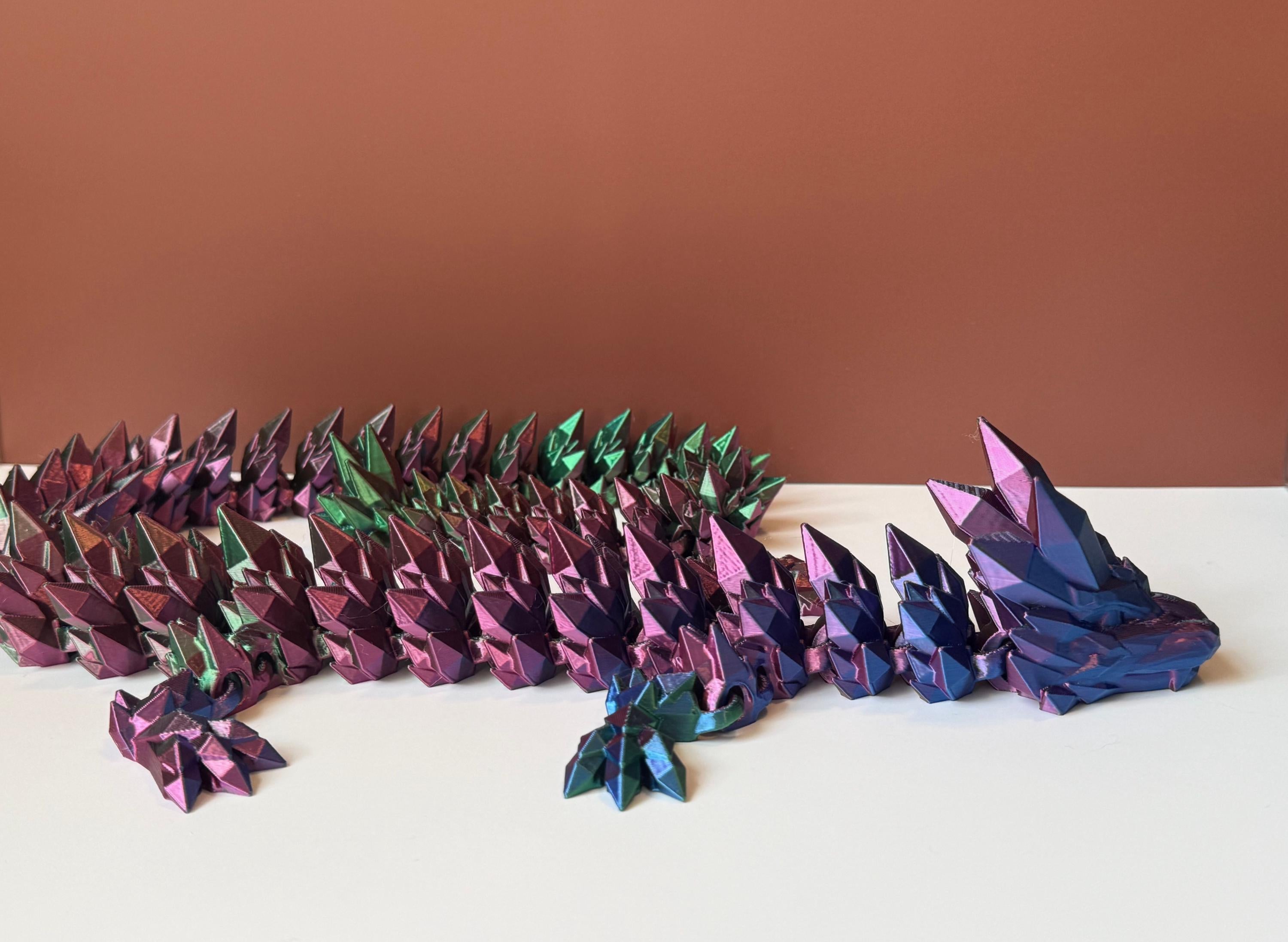 Multicolored 3D printed dragon with crystal-shaped features across the entire body, placed on a white surface with a brown backdrop