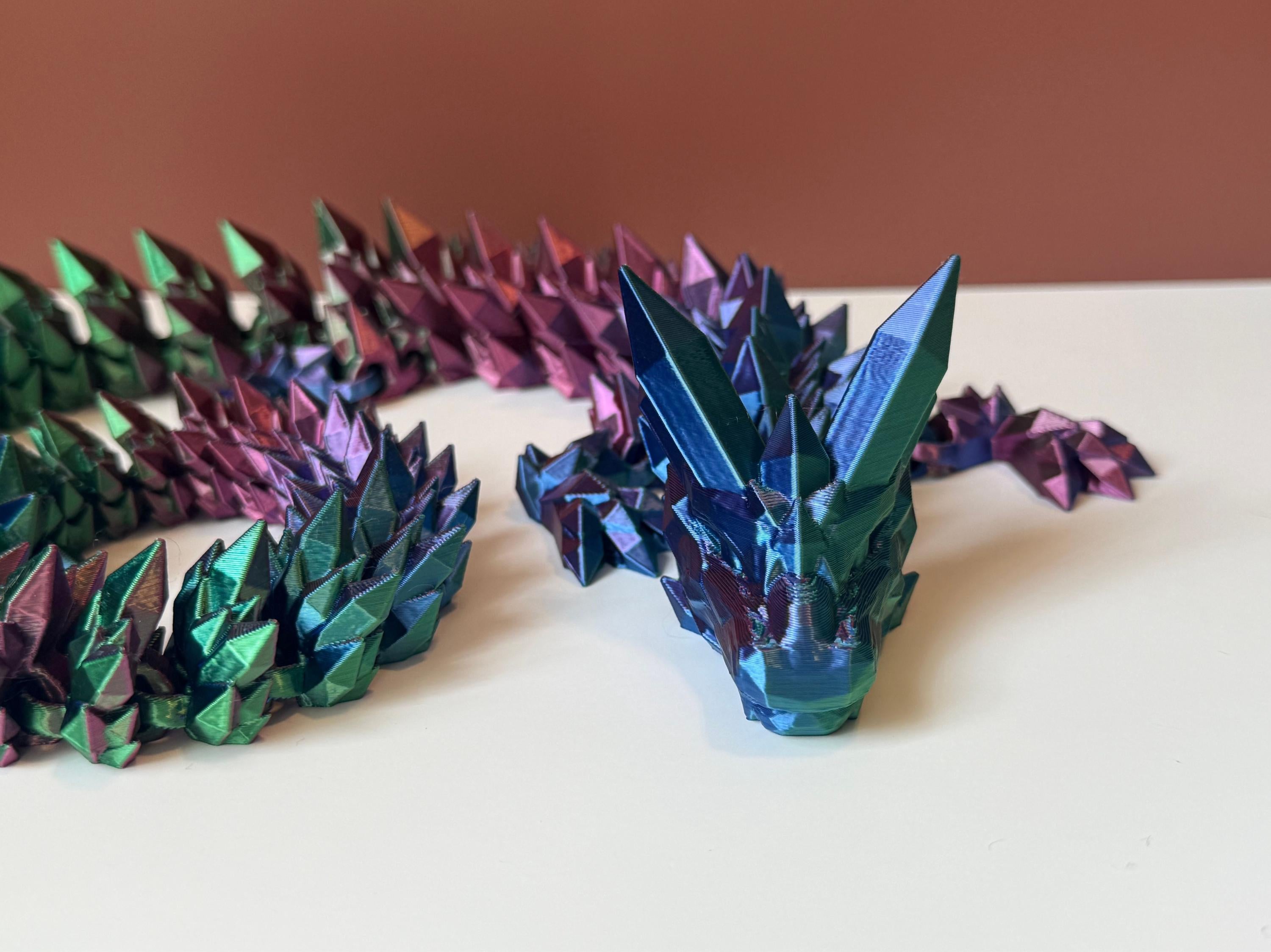 Multicolored 3D printed dragon with crystal-shaped features across the entire body, placed on a white surface with a brown backdrop