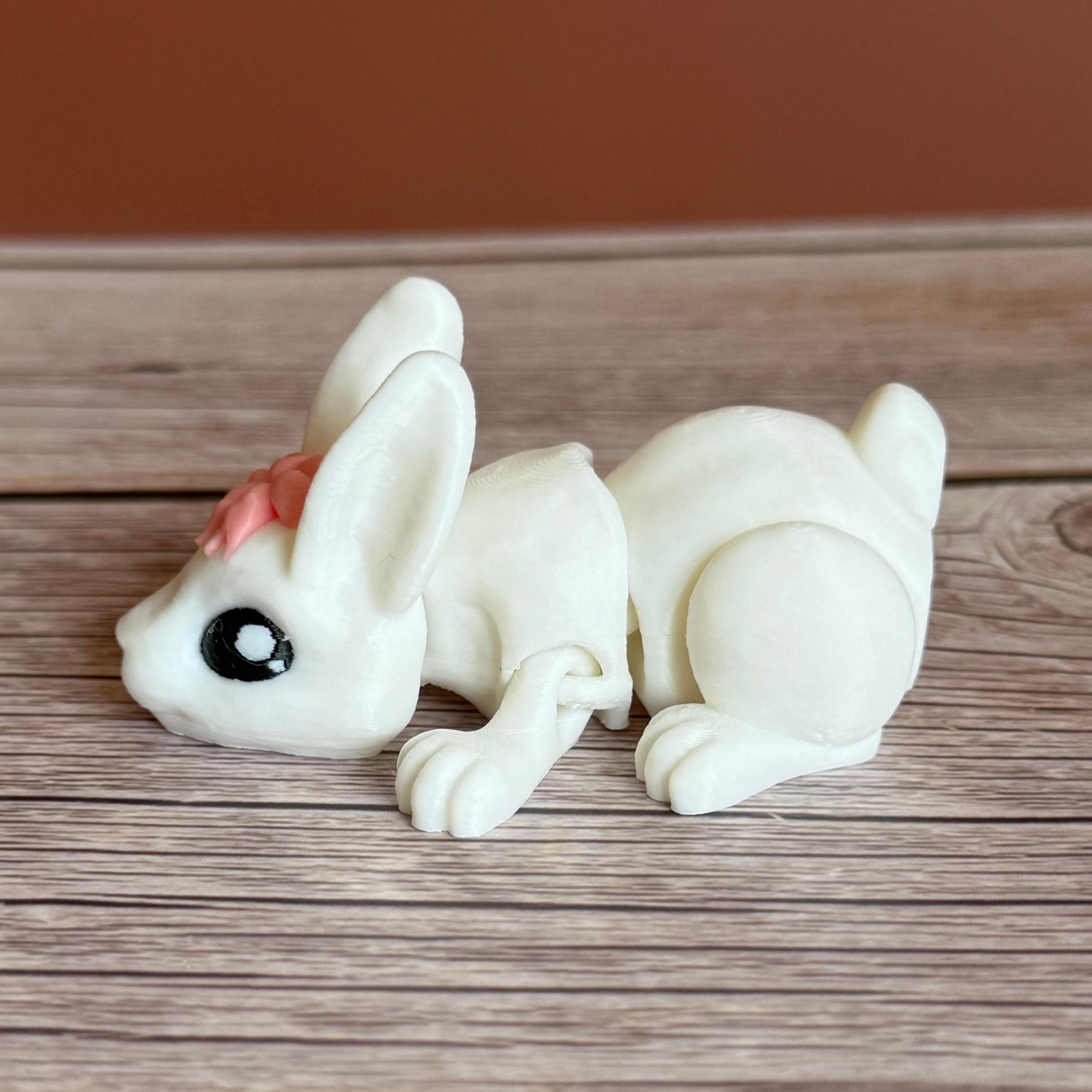 Side of a 3D printed white rabbit figurine with a pink bow on a wooden surface