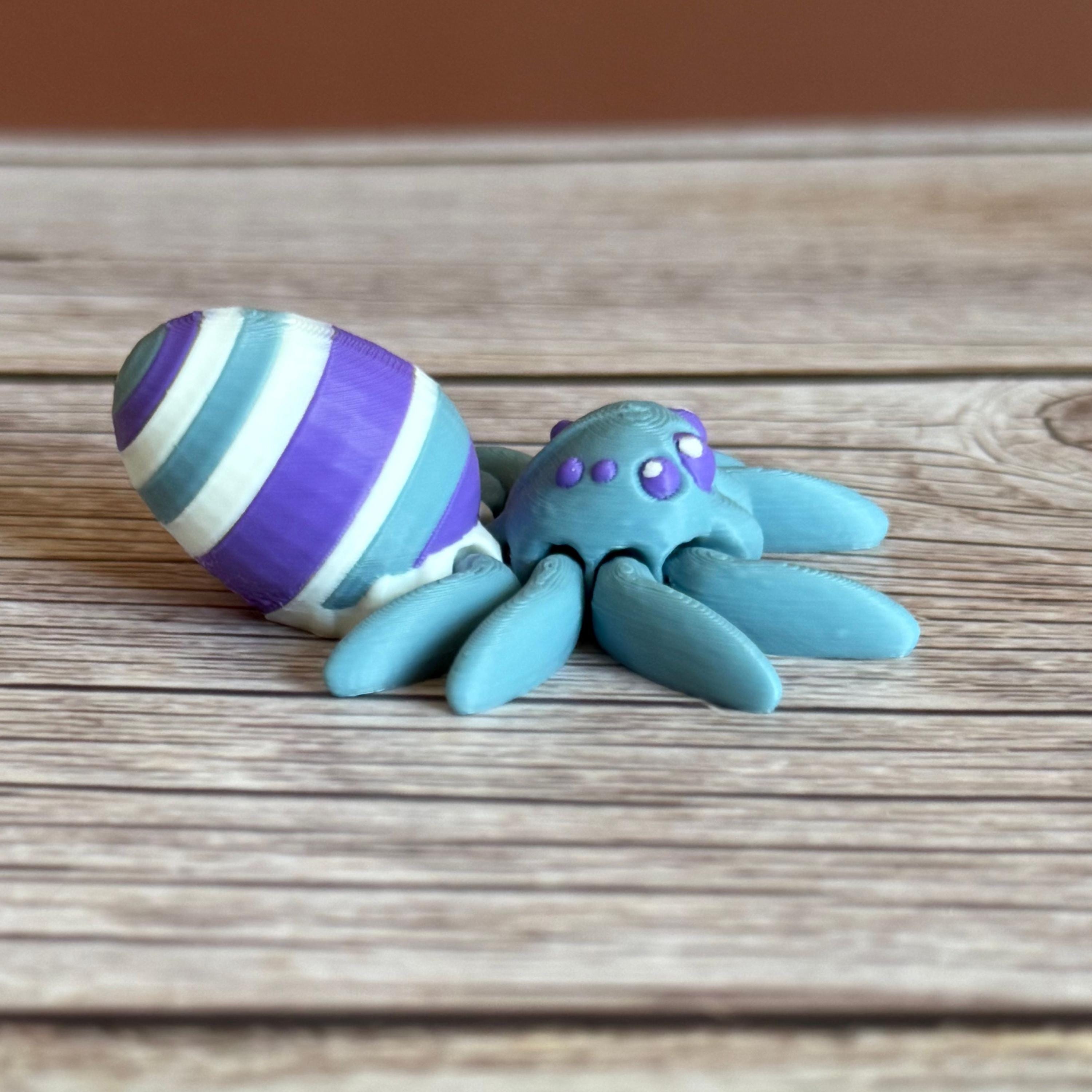 Side of a 3D printed blue spider featuring a white, blue, and purple egg on its body, placed on a wooden surface