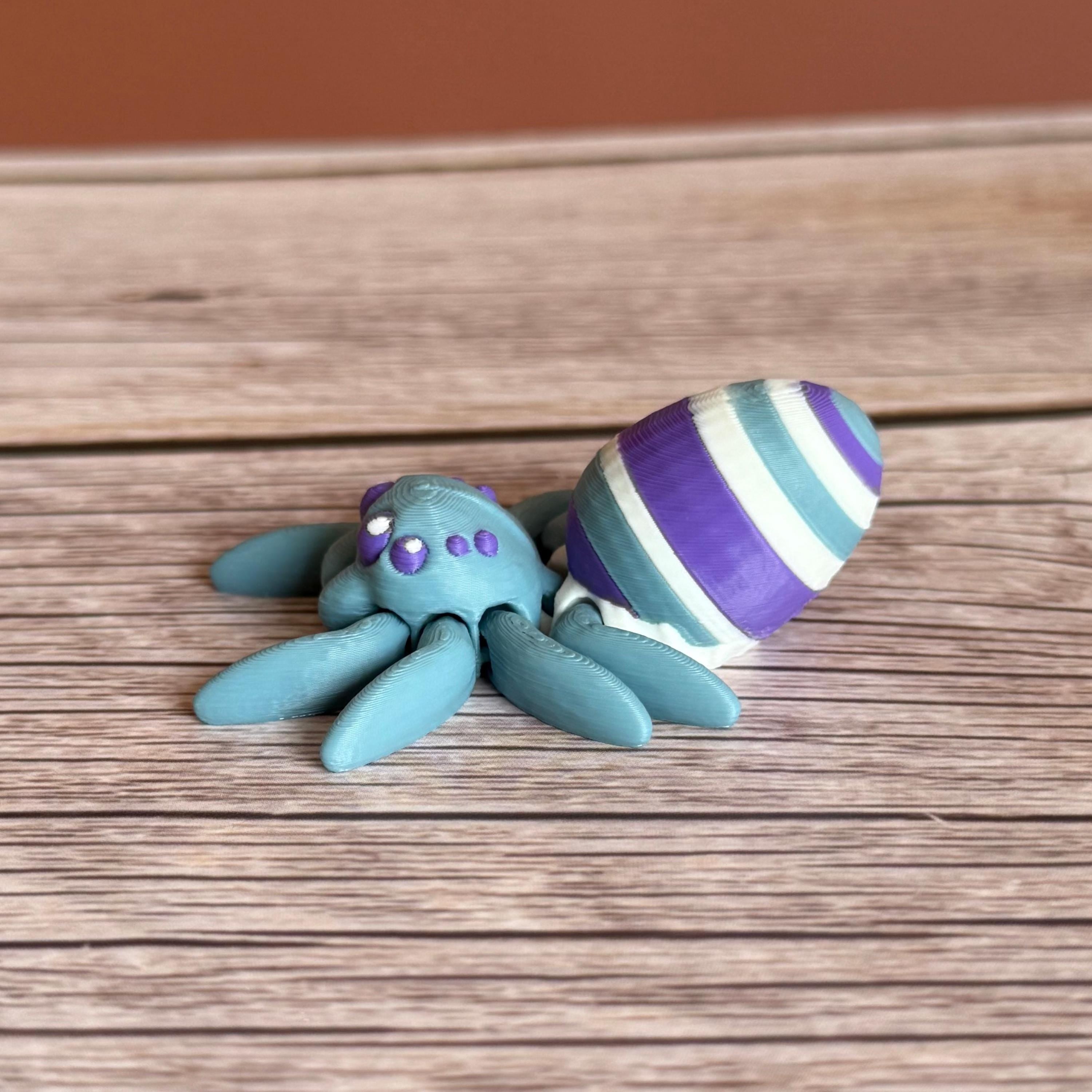 Side of a 3D printed blue spider featuring a white, blue, and purple egg on its body, placed on a wooden surface