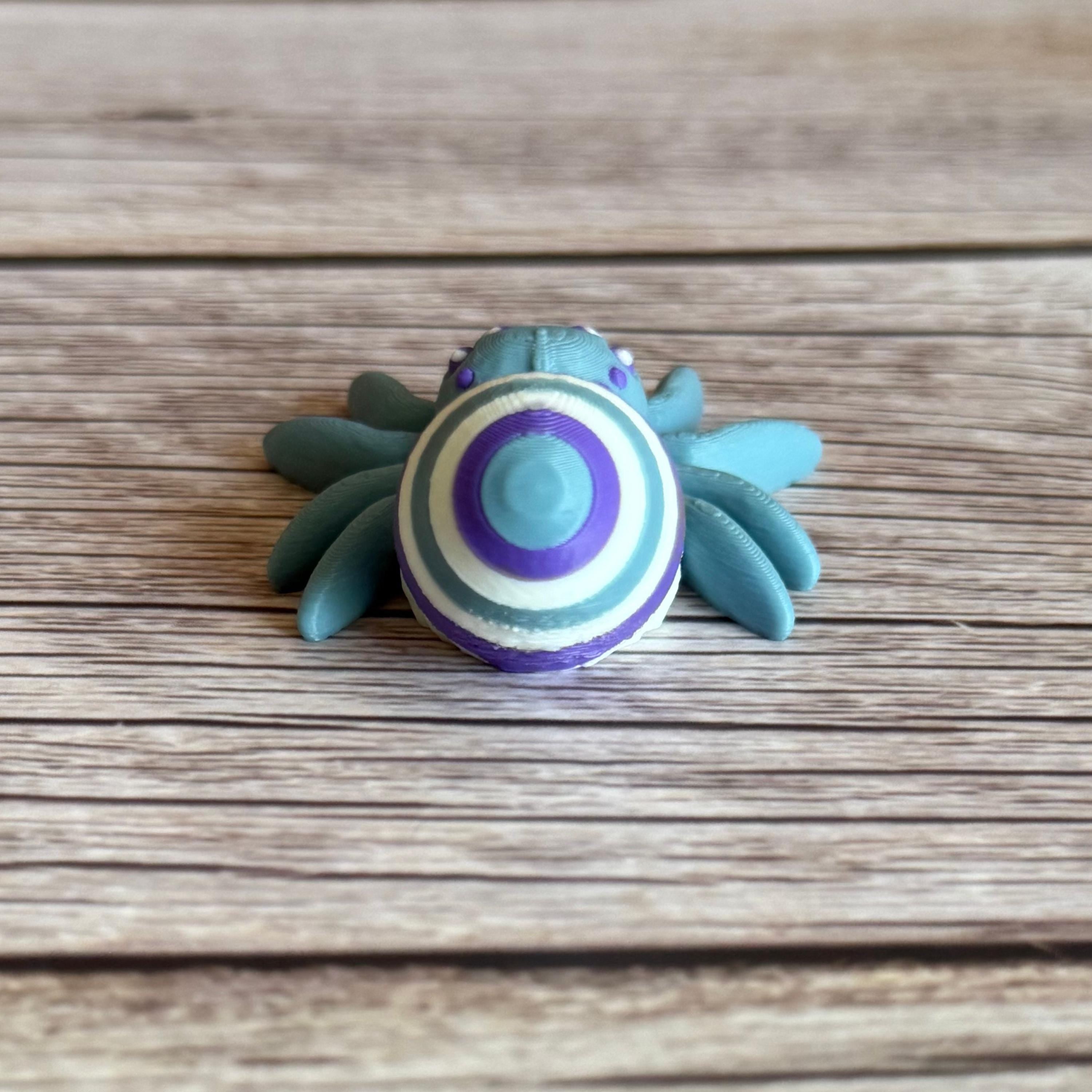 Back of a 3D printed blue spider featuring a white, blue, and purple egg on its body, placed on a wooden surface
