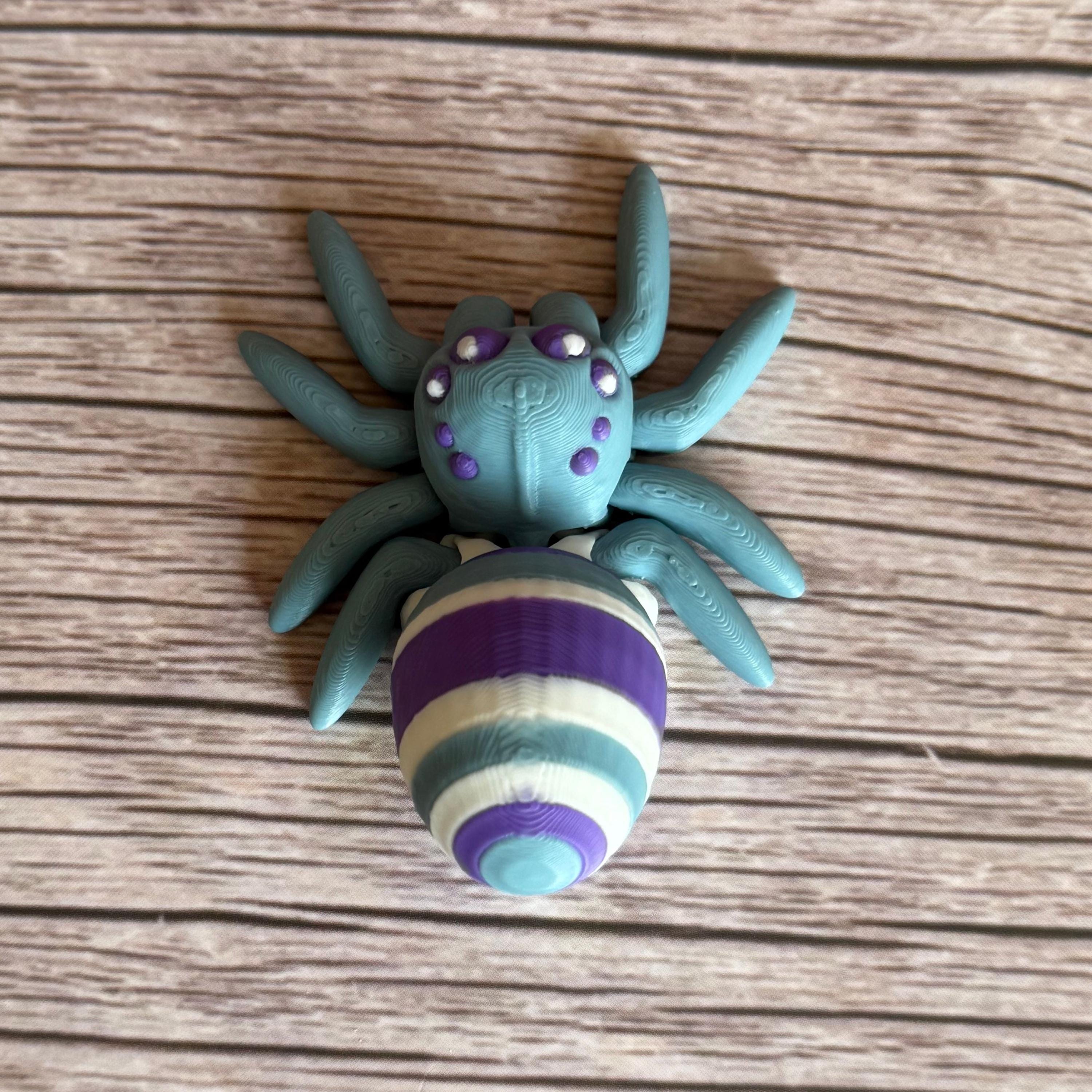 Top of a 3D printed blue spider featuring a white, blue, and purple egg on its body, placed on a wooden surface