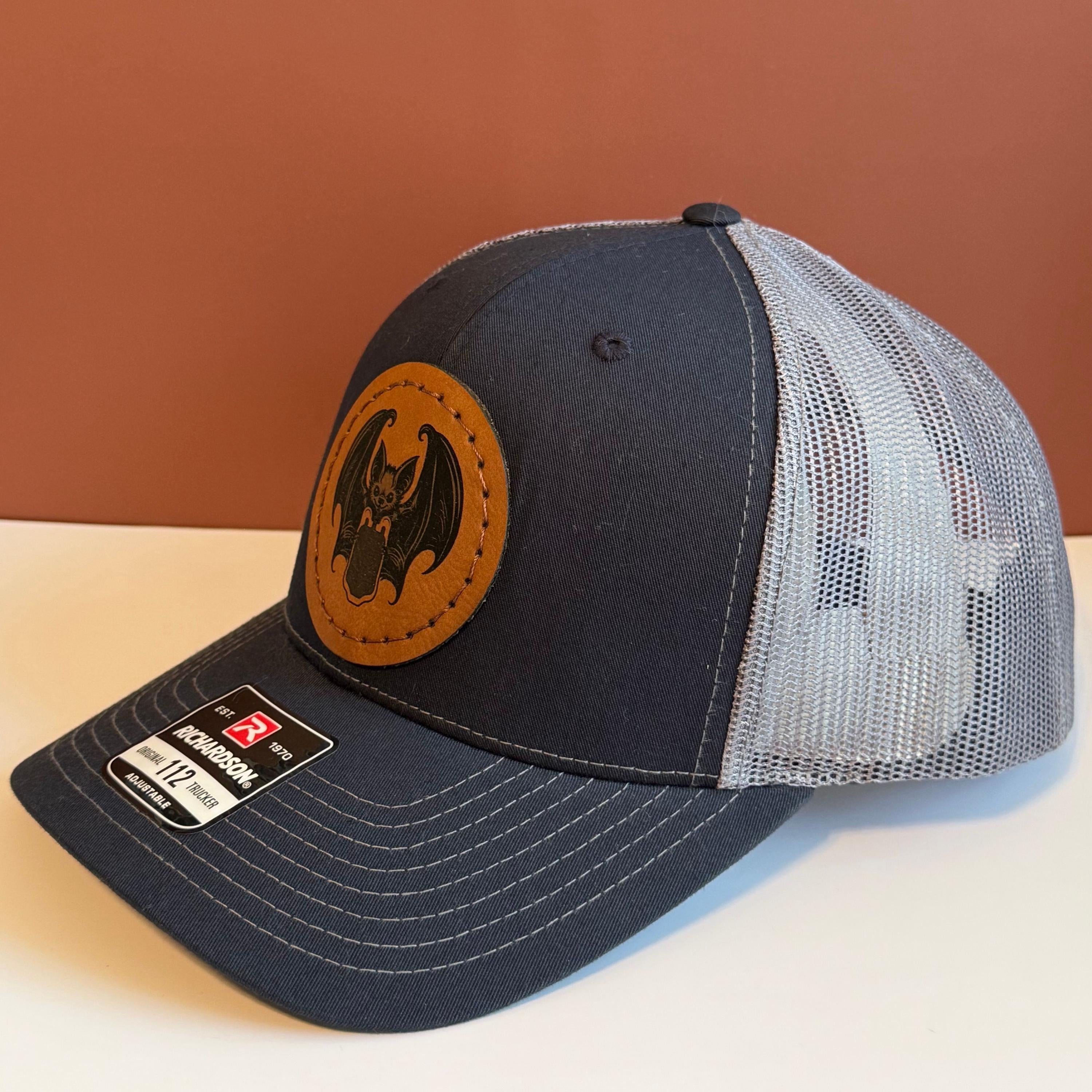 A blue and gray snapback hat with a hand-sewn leatherette patch featuring a vampire bat holding a blood bag, placed against a white and brown background