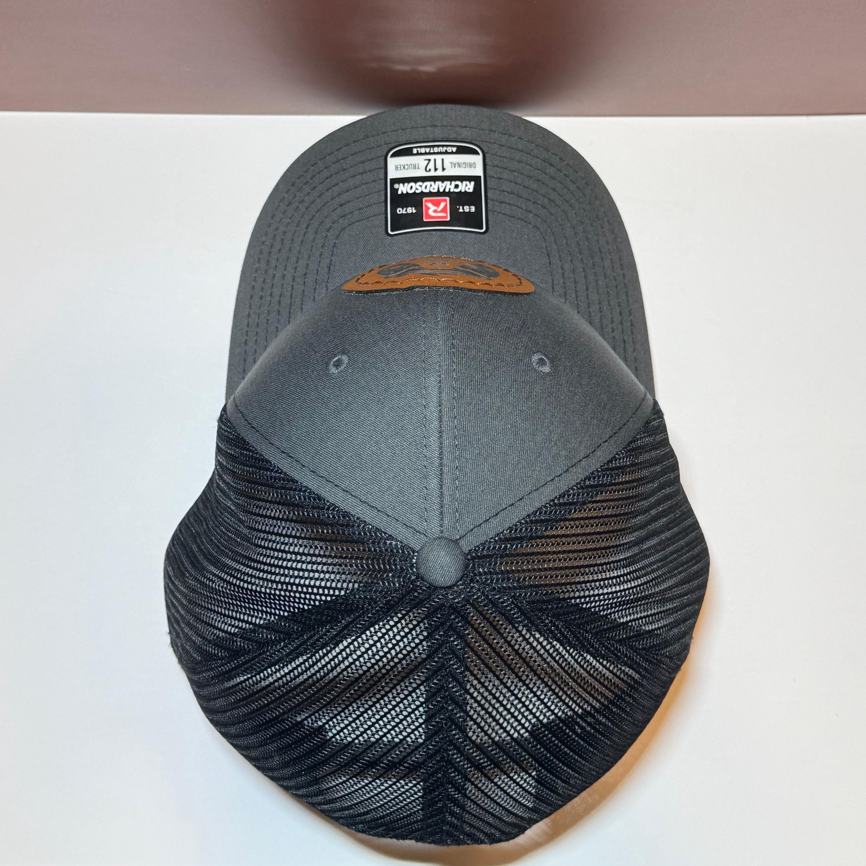 Top of gray and black mesh-back trucker hat with Richardson 112 sticker and patch on a white and brown background.