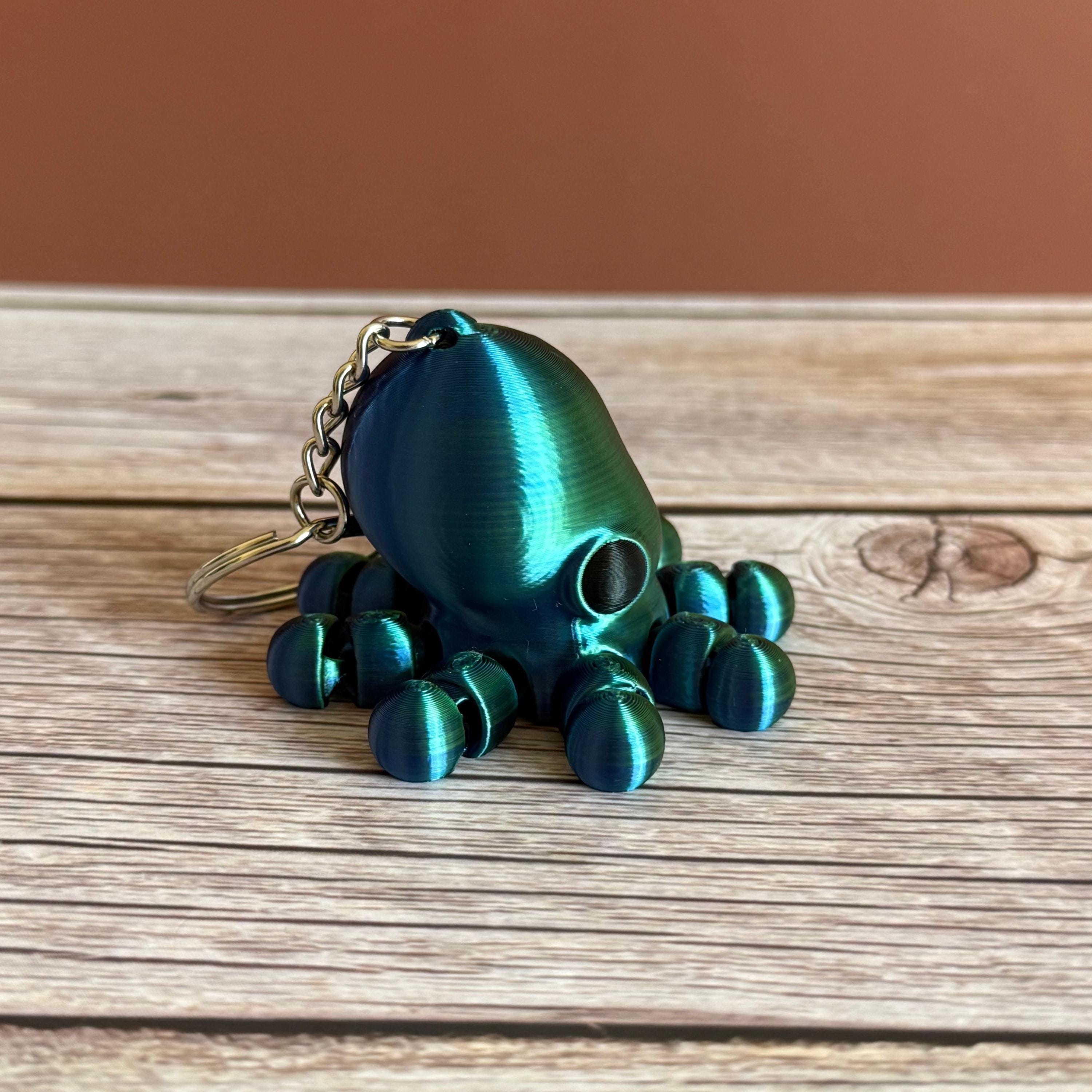 3D printed shiny green and blue octopus-shaped keychain, placed on a wooden surface with a brown background