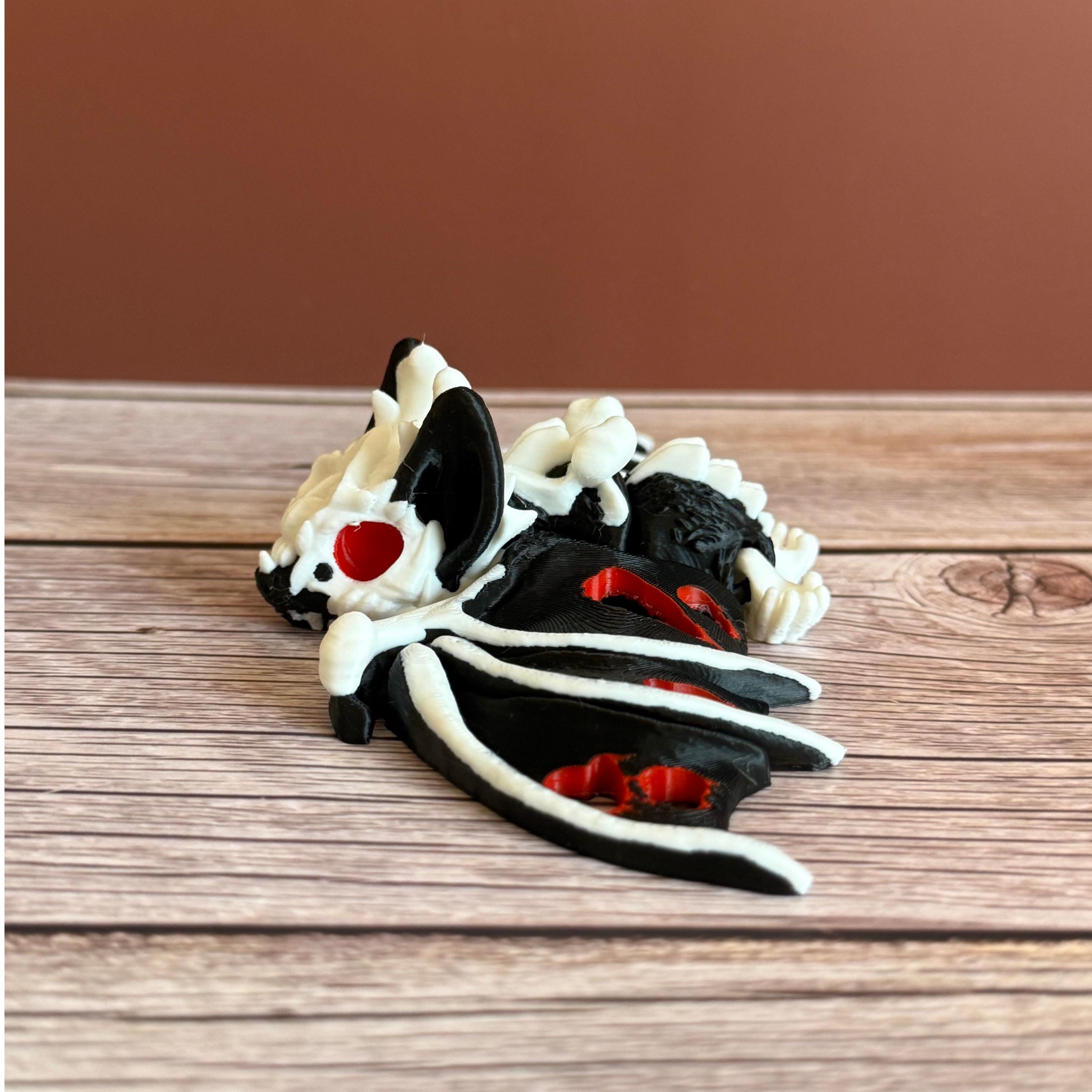 Side of a 3D printed skeleton-like bat with bloody appearing wings and red eyes, placed on a wooden surface and brown background