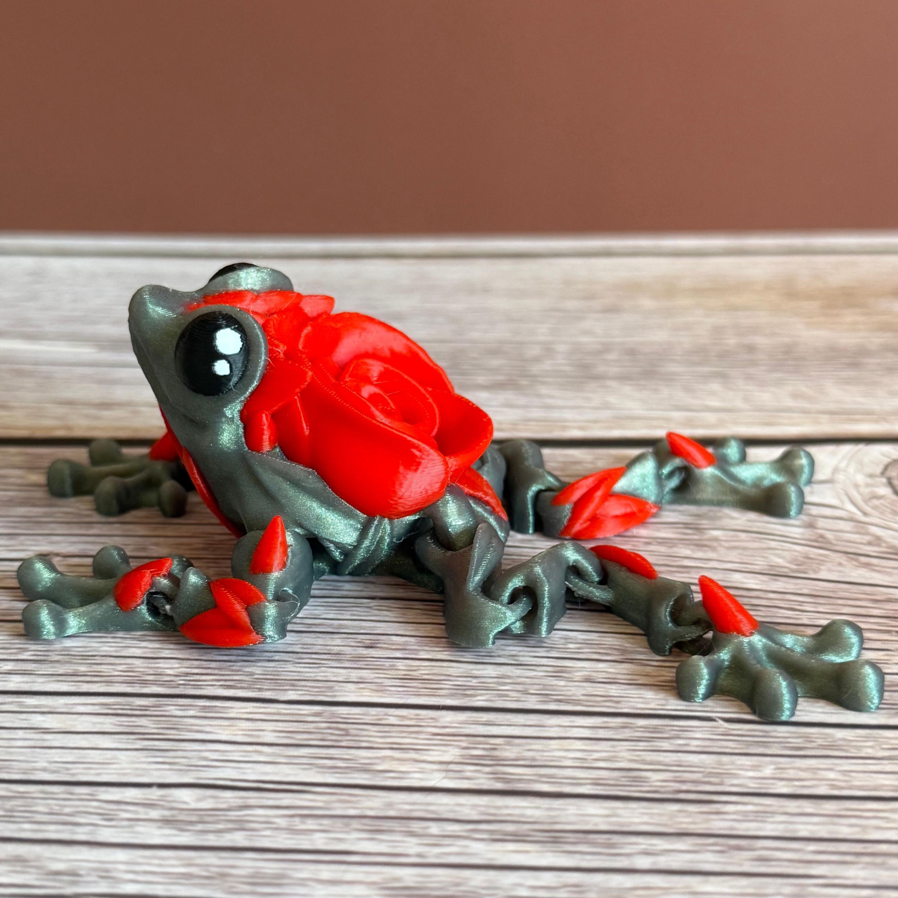Side of a 3D printed shimmer-green frog with red rose on it's back, placed on a wooden surface against a brown background