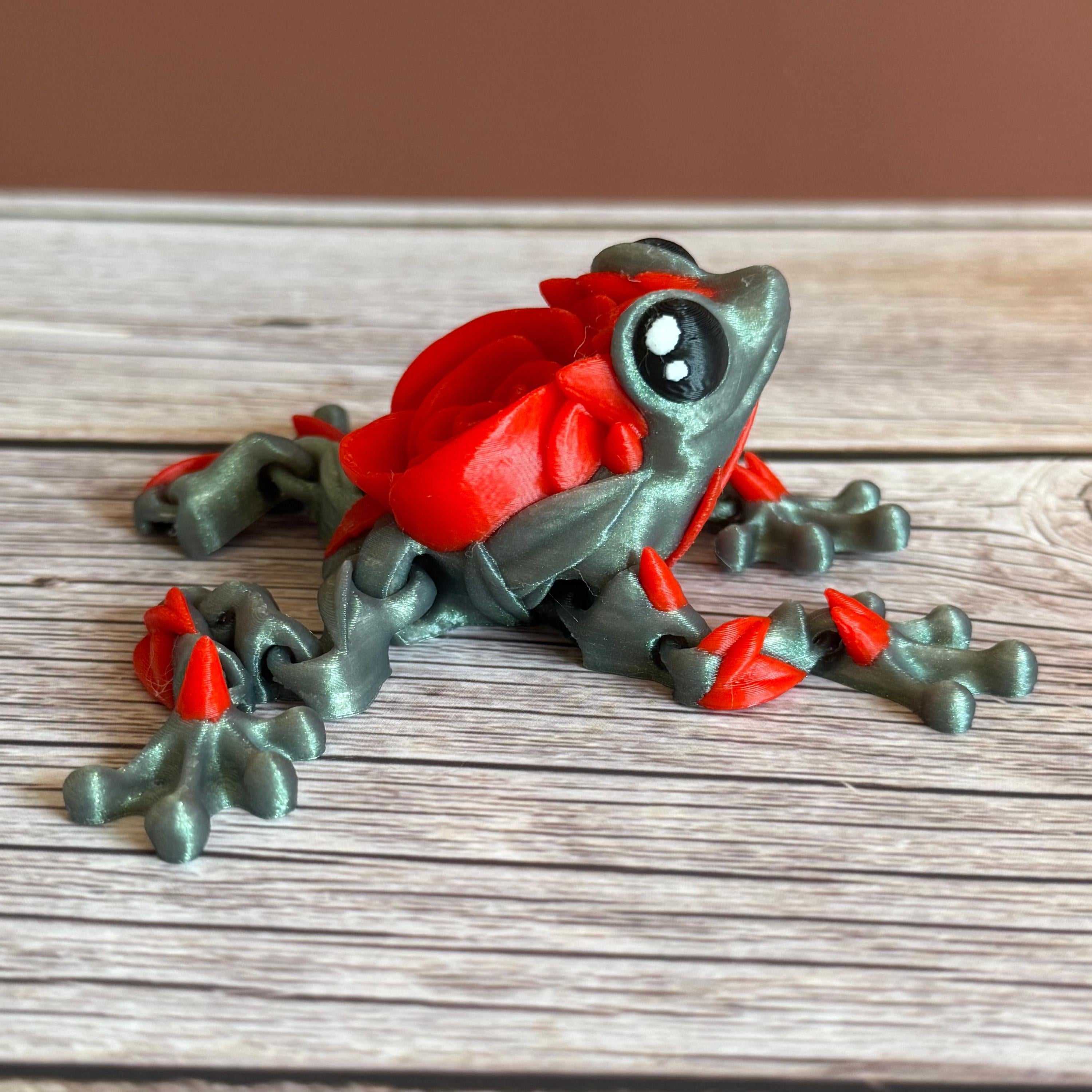 Side of a 3D printed shimmer-green frog with red rose on it's back, placed on a wooden surface against a brown background