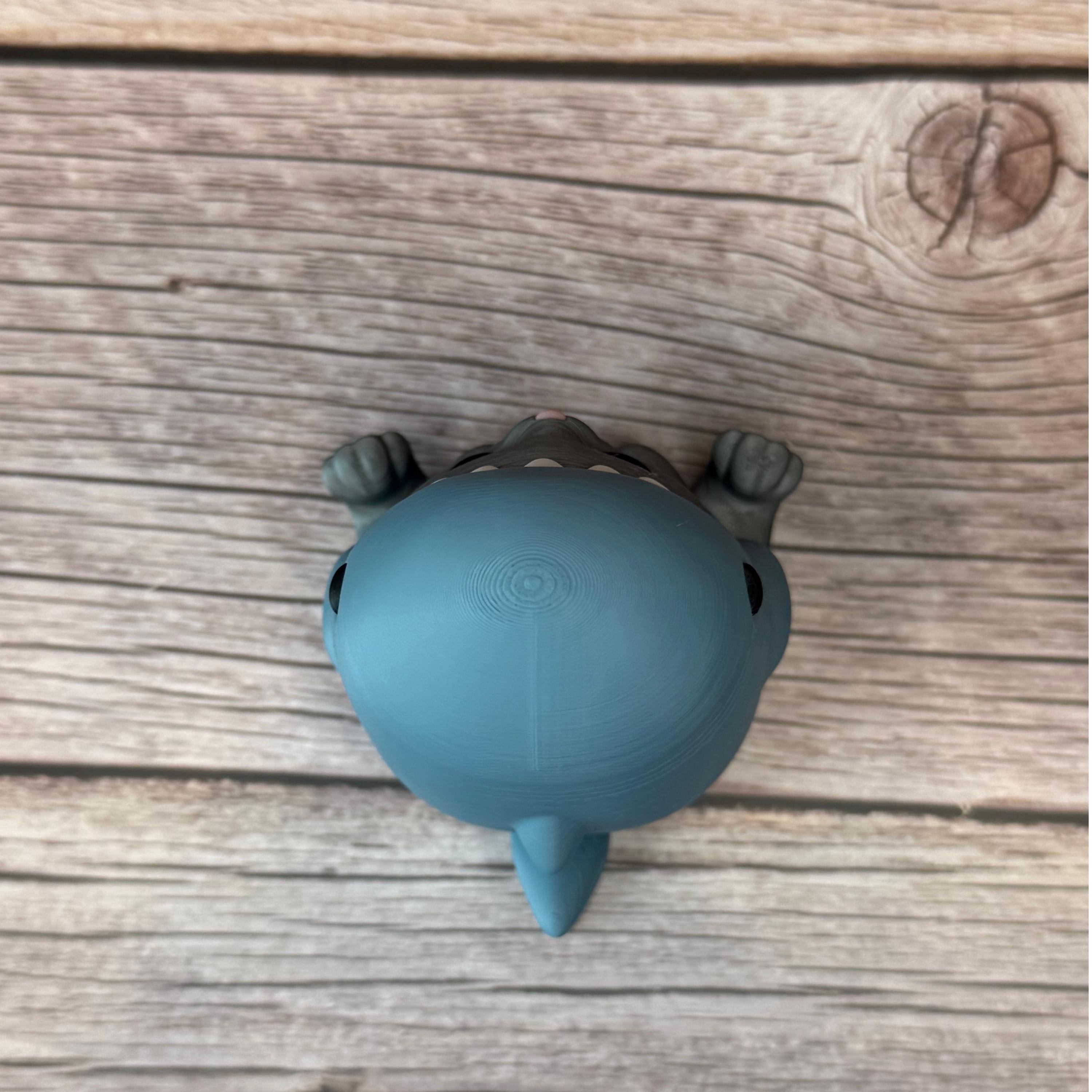 3D printed figurine of a gray cat wearing a shark costume in blue-gray color, placed on a wooden surface