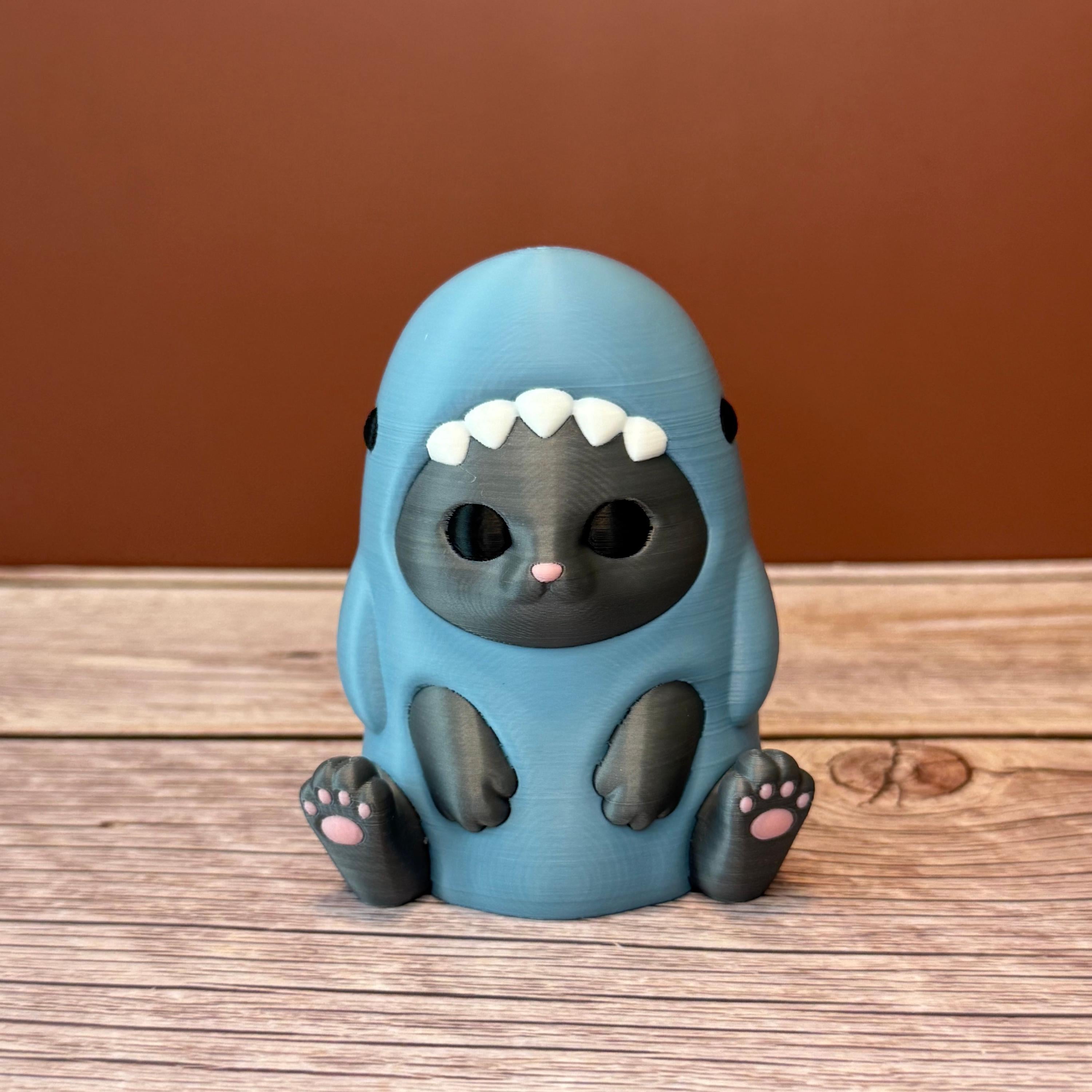 3D printed figurine of a gray cat wearing a shark costume in blue-gray color, placed on a wooden surface
