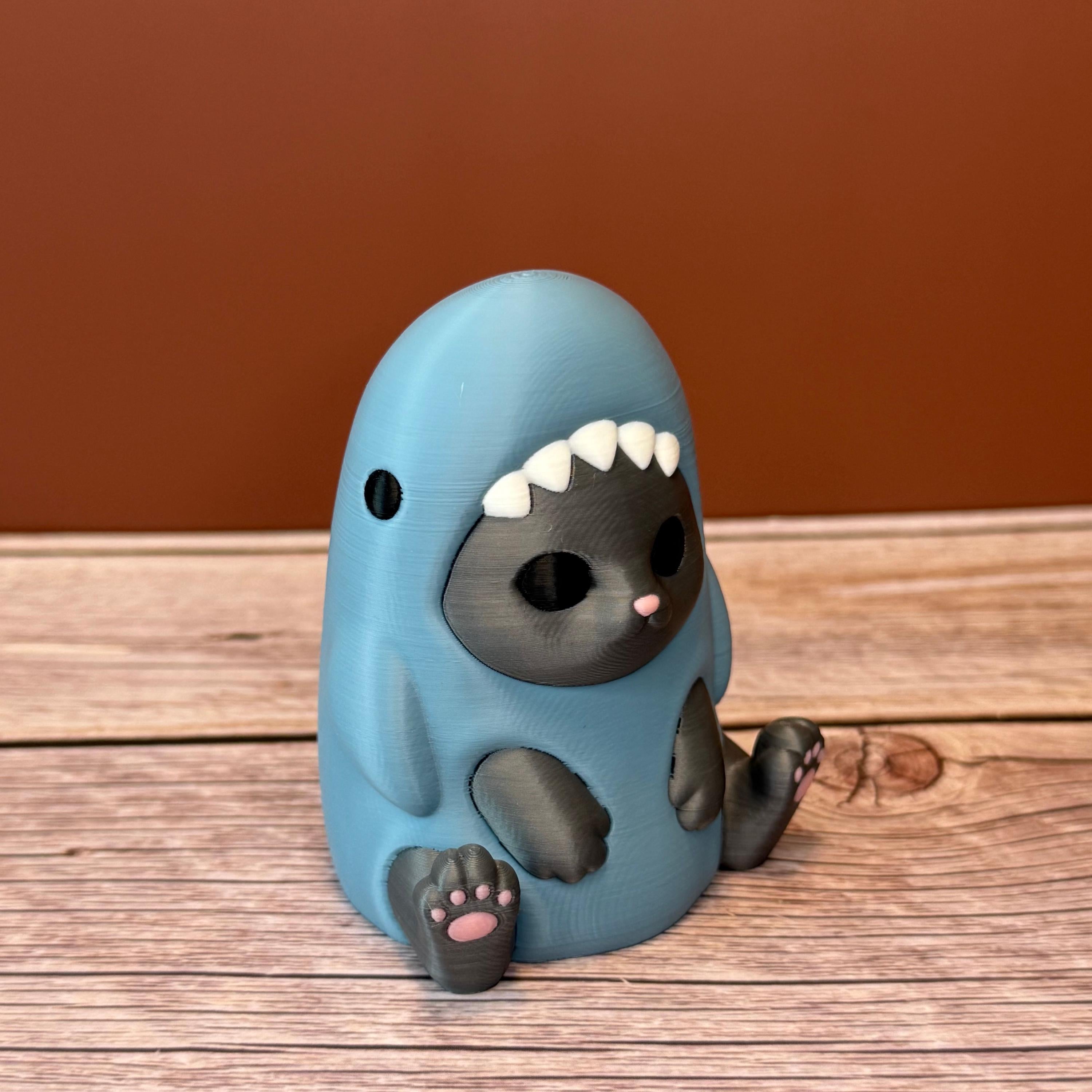 3D printed figurine of a gray cat wearing a shark costume in blue-gray color, placed on a wooden surface