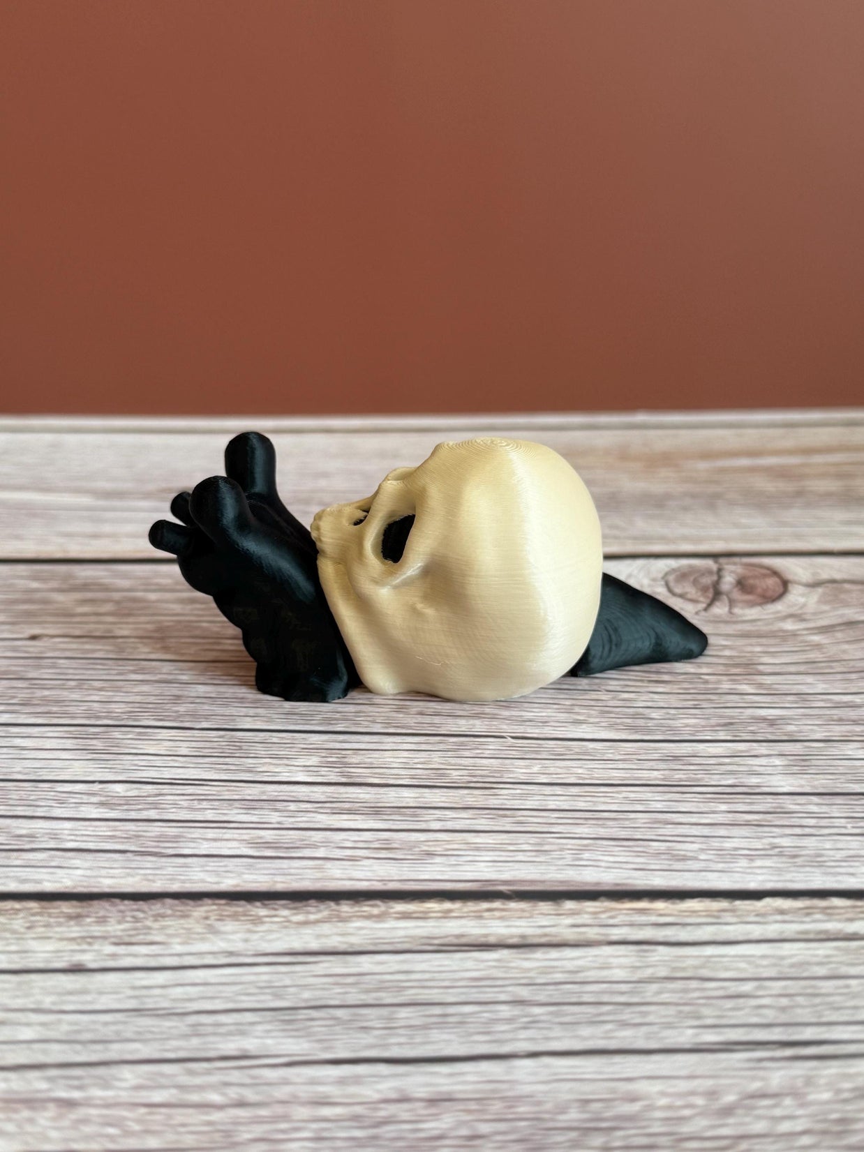 Black 3D printed articulated snail with a beige shell shaped like a human skull, placed on a wooden surface