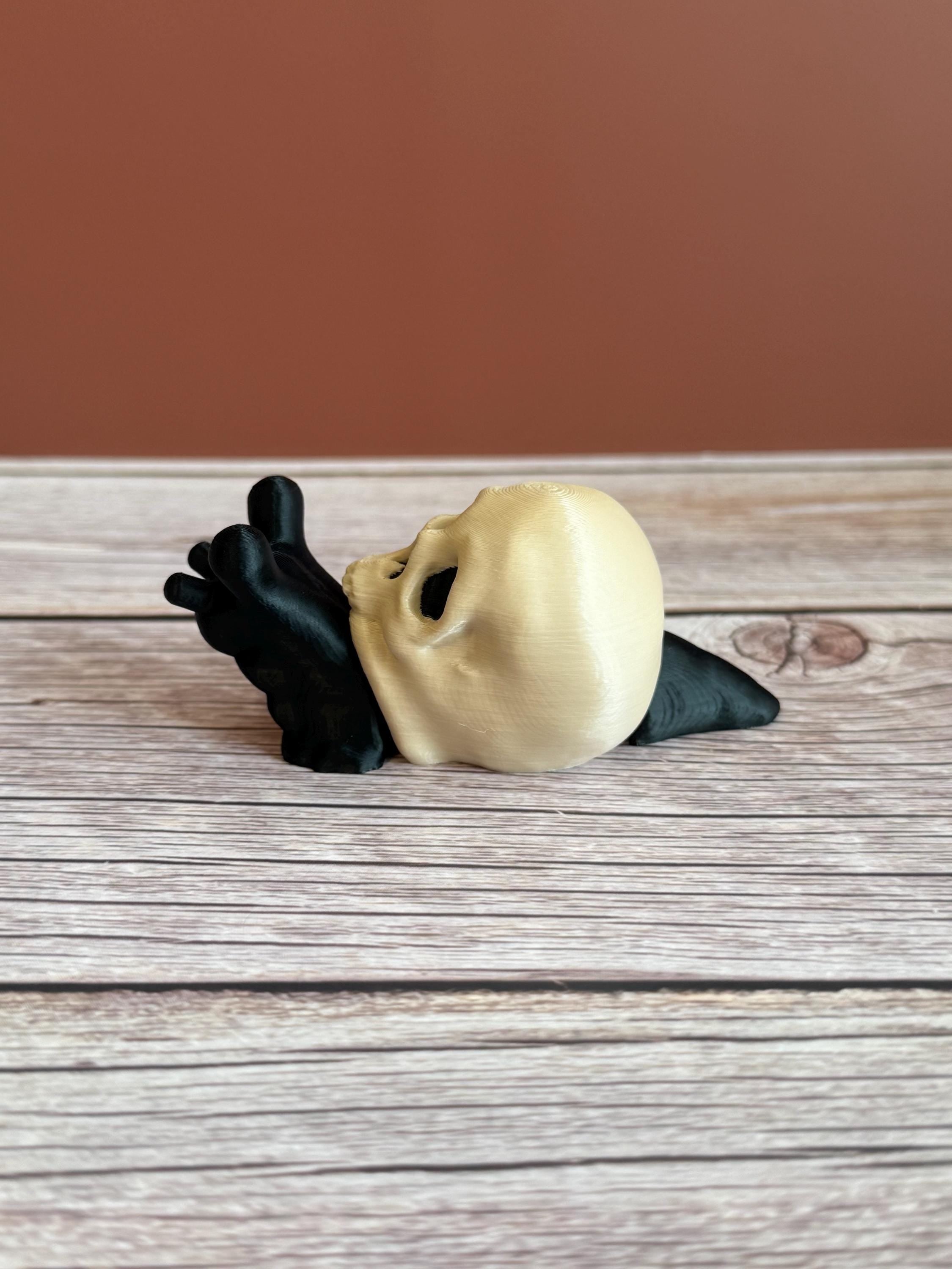 Black 3D printed articulated snail with a beige shell shaped like a human skull, placed on a wooden surface