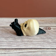 Black 3D printed articulated snail with a beige shell shaped like a human skull, placed on a wooden surface