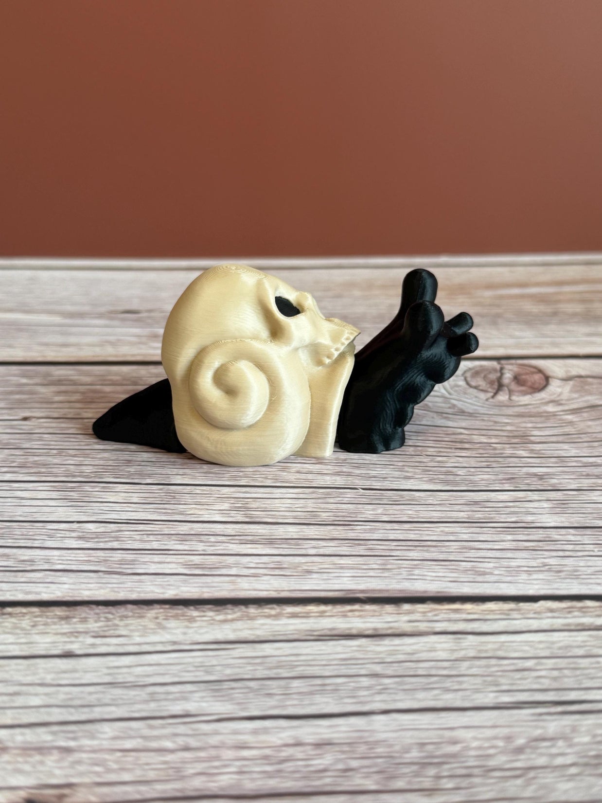 Black 3D printed articulated snail with a beige shell shaped like a human skull, placed on a wooden surface