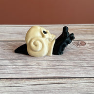 Black 3D printed articulated snail with a beige shell shaped like a human skull, placed on a wooden surface