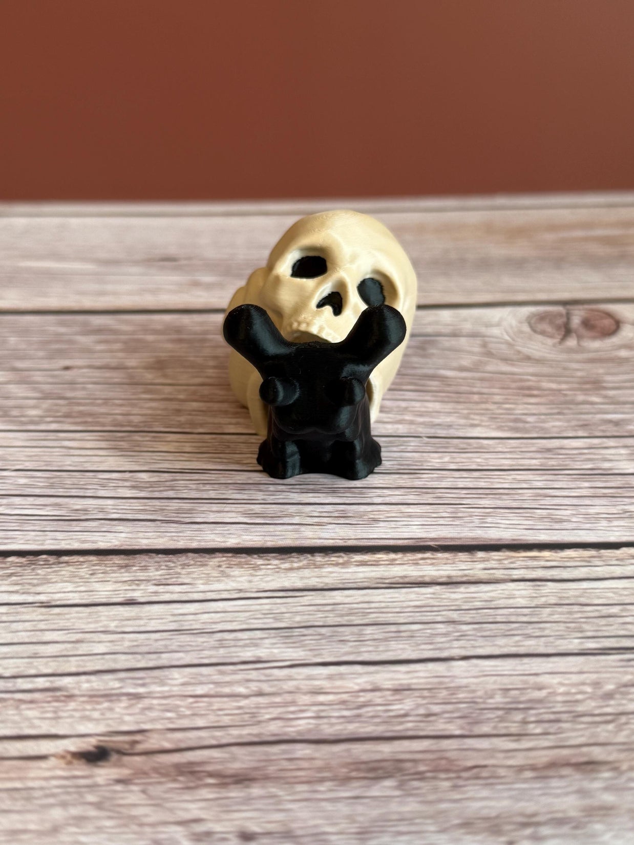 Black 3D printed articulated snail with a beige shell shaped like a human skull, placed on a wooden surface