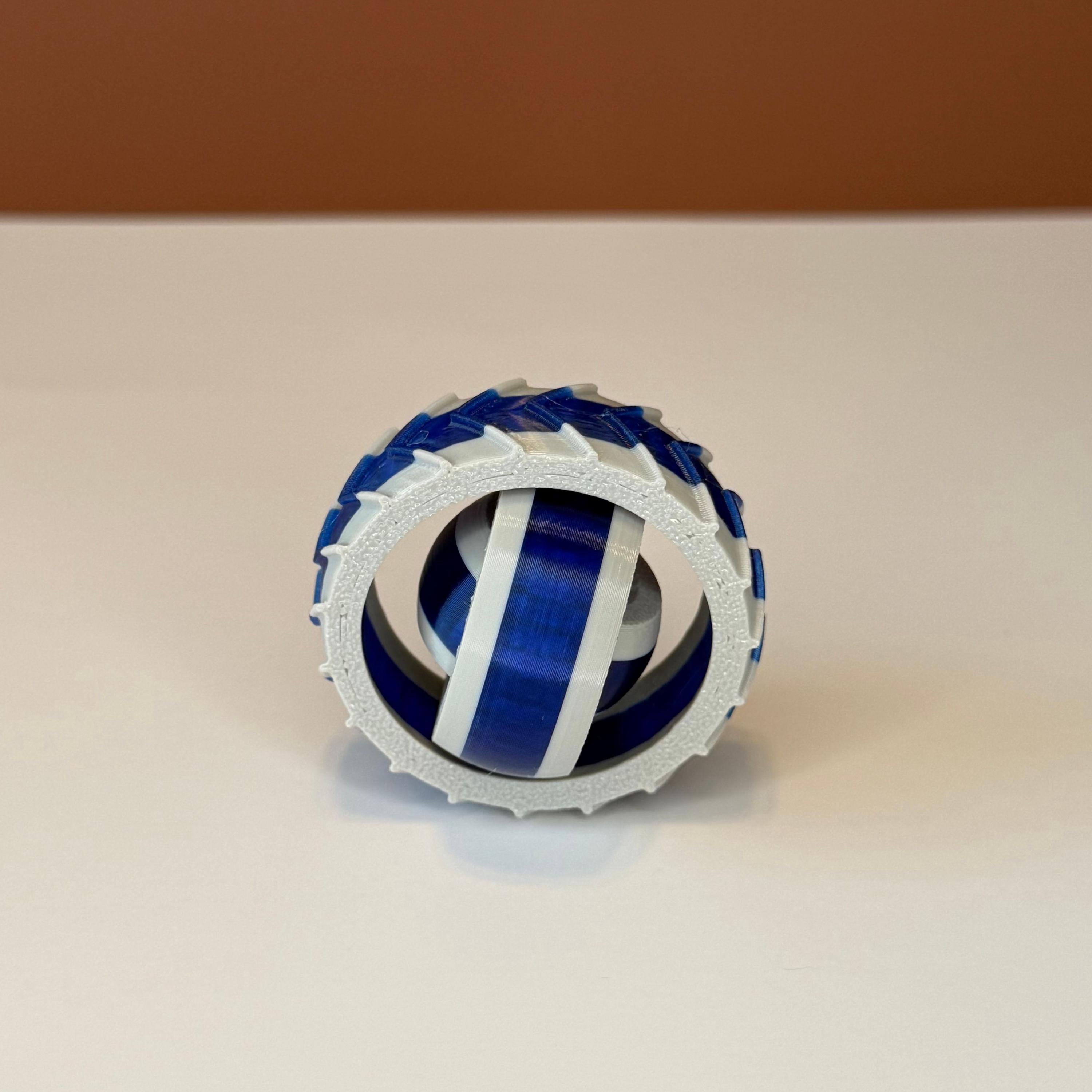 3D printed gray and blue gyro spinner consisting of 3 rings set into each other that spin in different directions on a white and brown background