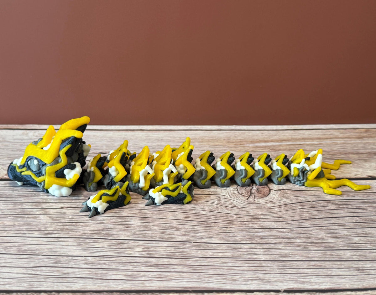 A 3D printed, articulated toy dragon figurine with yellow and black coloring, featuring lightning bolt details and storm cloud patterns, placed on a wooden surface