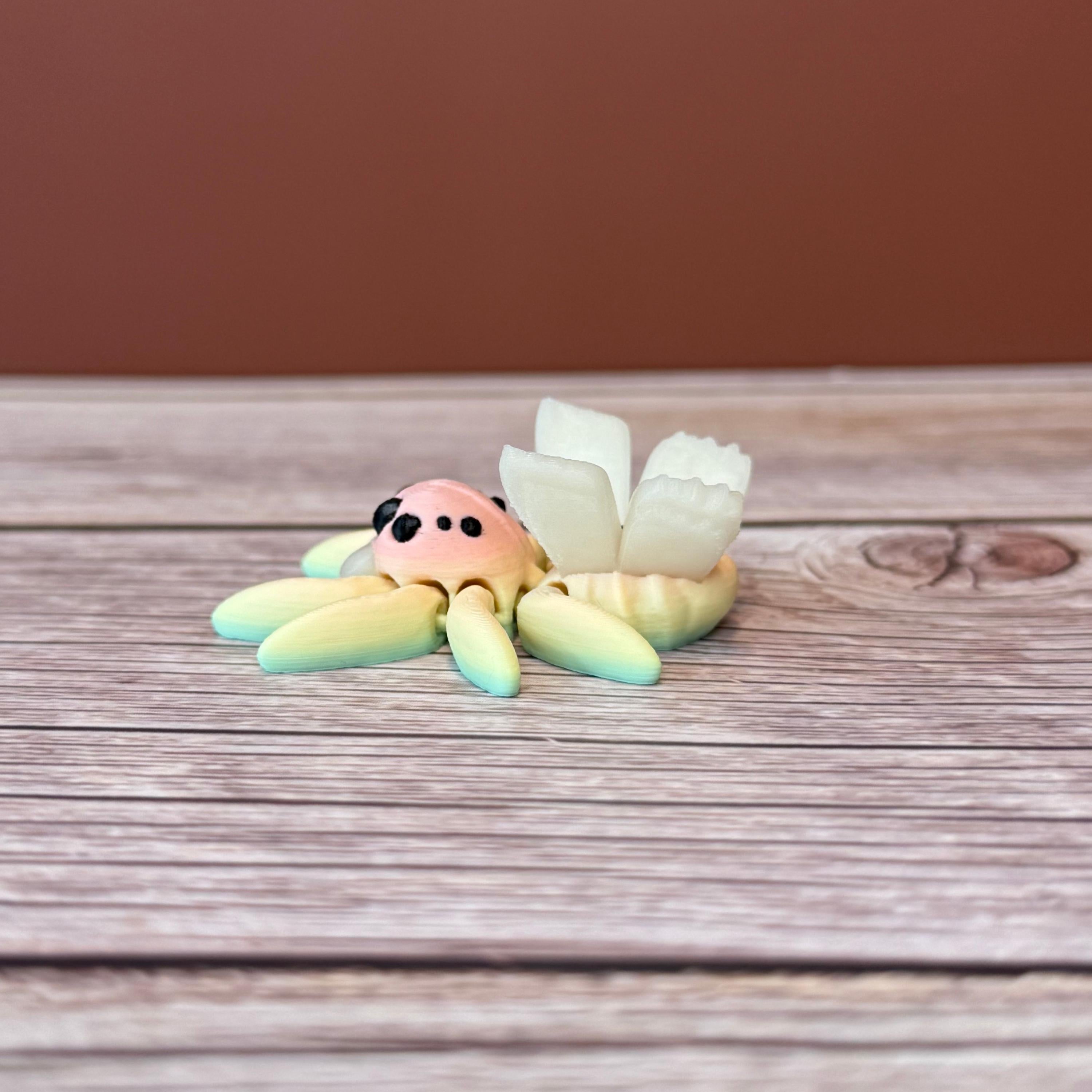 3D printed small spider with fairy design featuring rainbow coloring and white wings, placed on a wooden surface