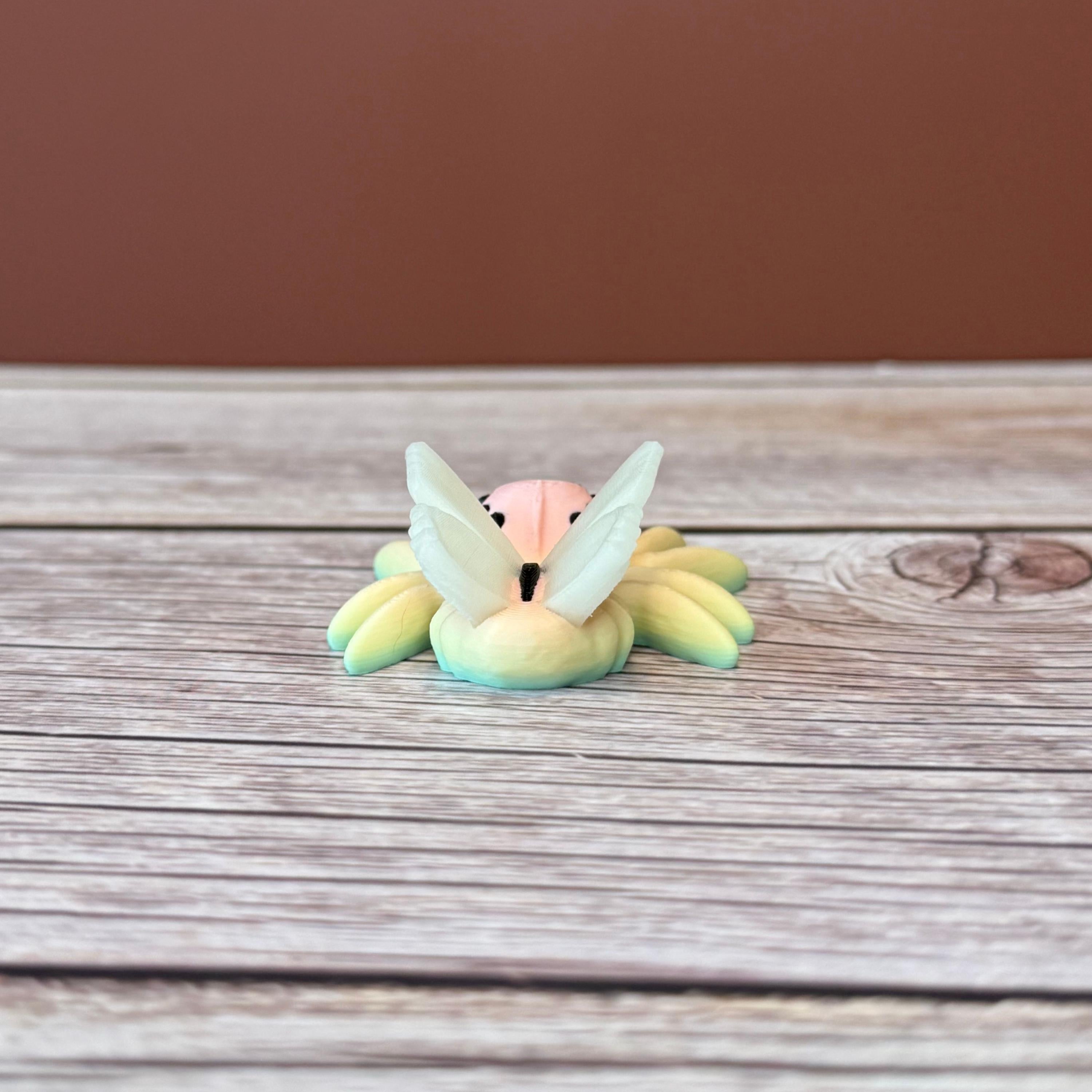 3D printed small spider with fairy design featuring rainbow coloring and white wings, placed on a wooden surface