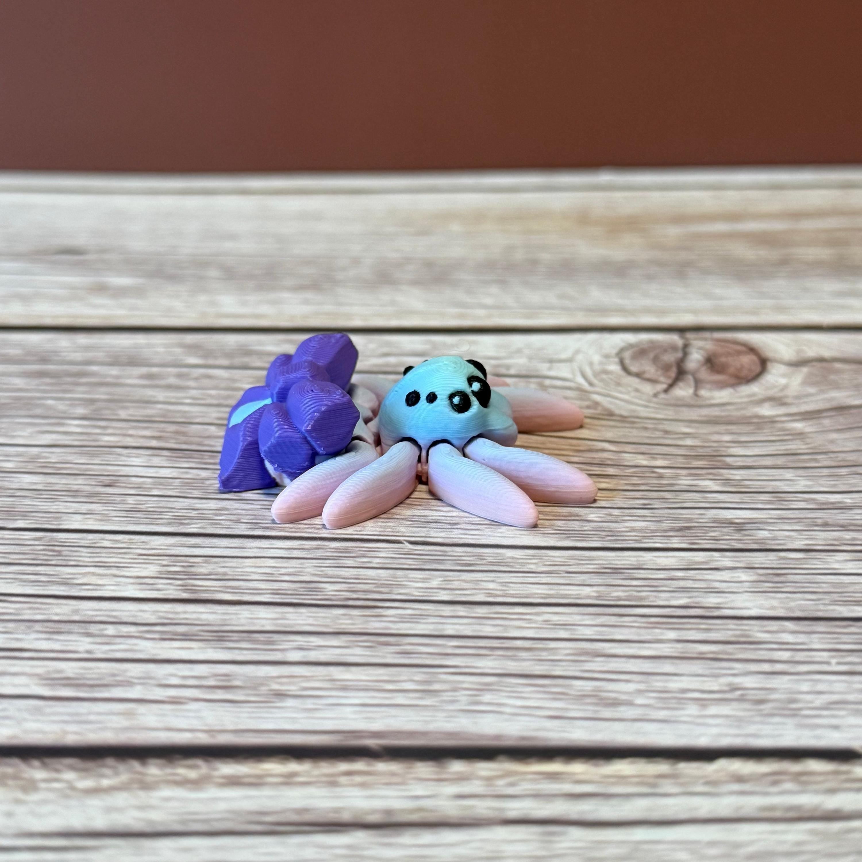A 3D printed toy figurine of an articulated spider with a giant purple bow and multicolored details on a wooden surface.