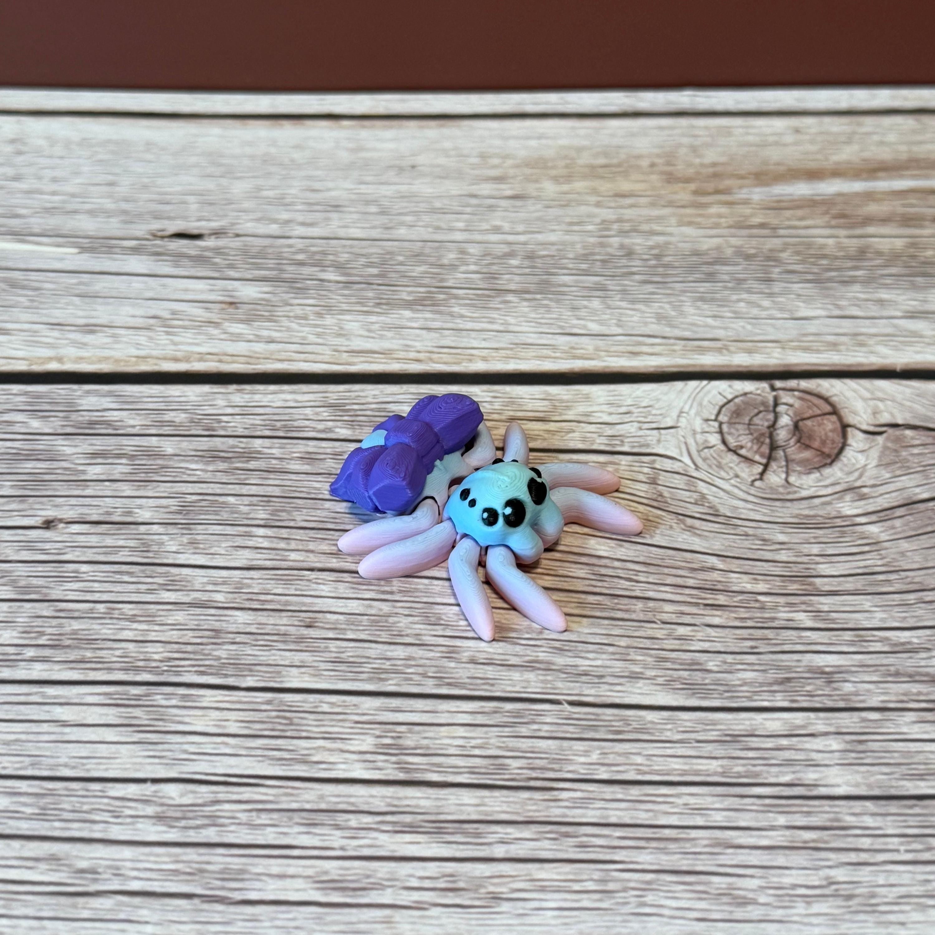 A 3D printed toy figurine of an articulated spider with a giant purple bow and multicolored details on a wooden surface.