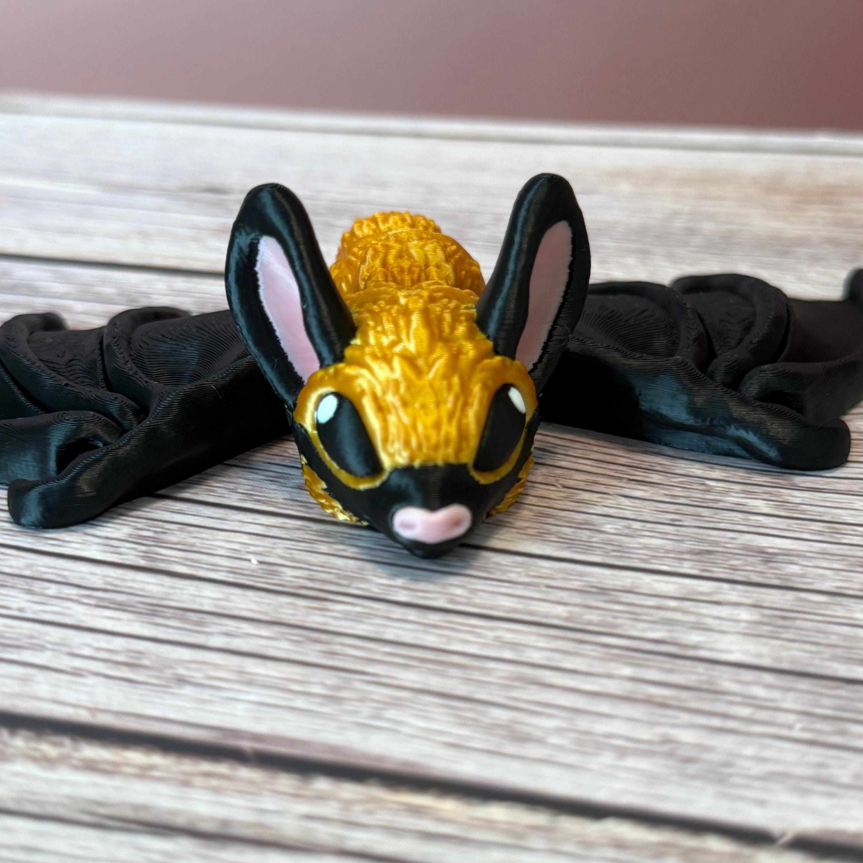3D printed gold and black articulated bat with tall pink ears and pink nose, placed on a wooden surface