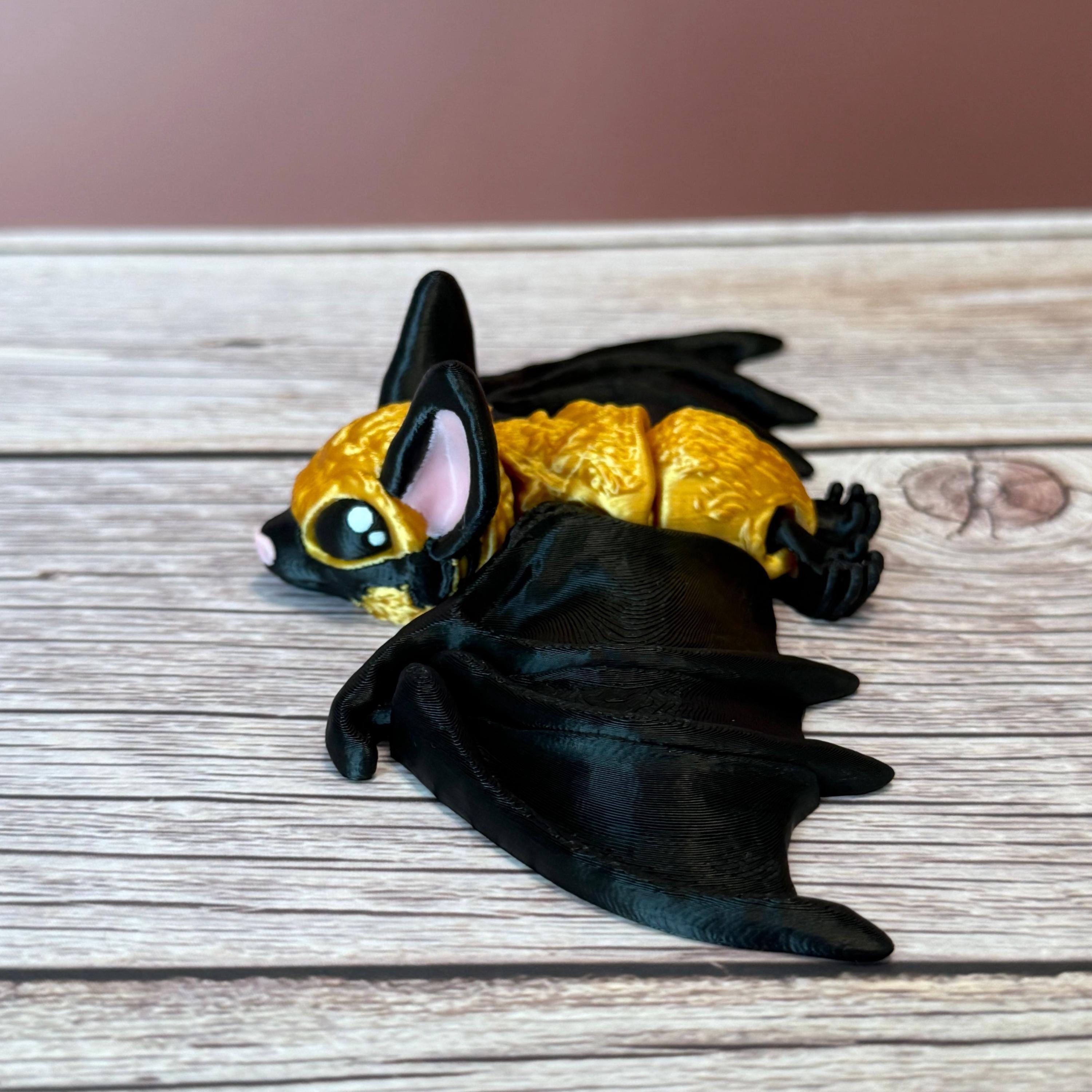 3D printed gold and black articulated bat with tall pink ears and pink nose, placed on a wooden surface